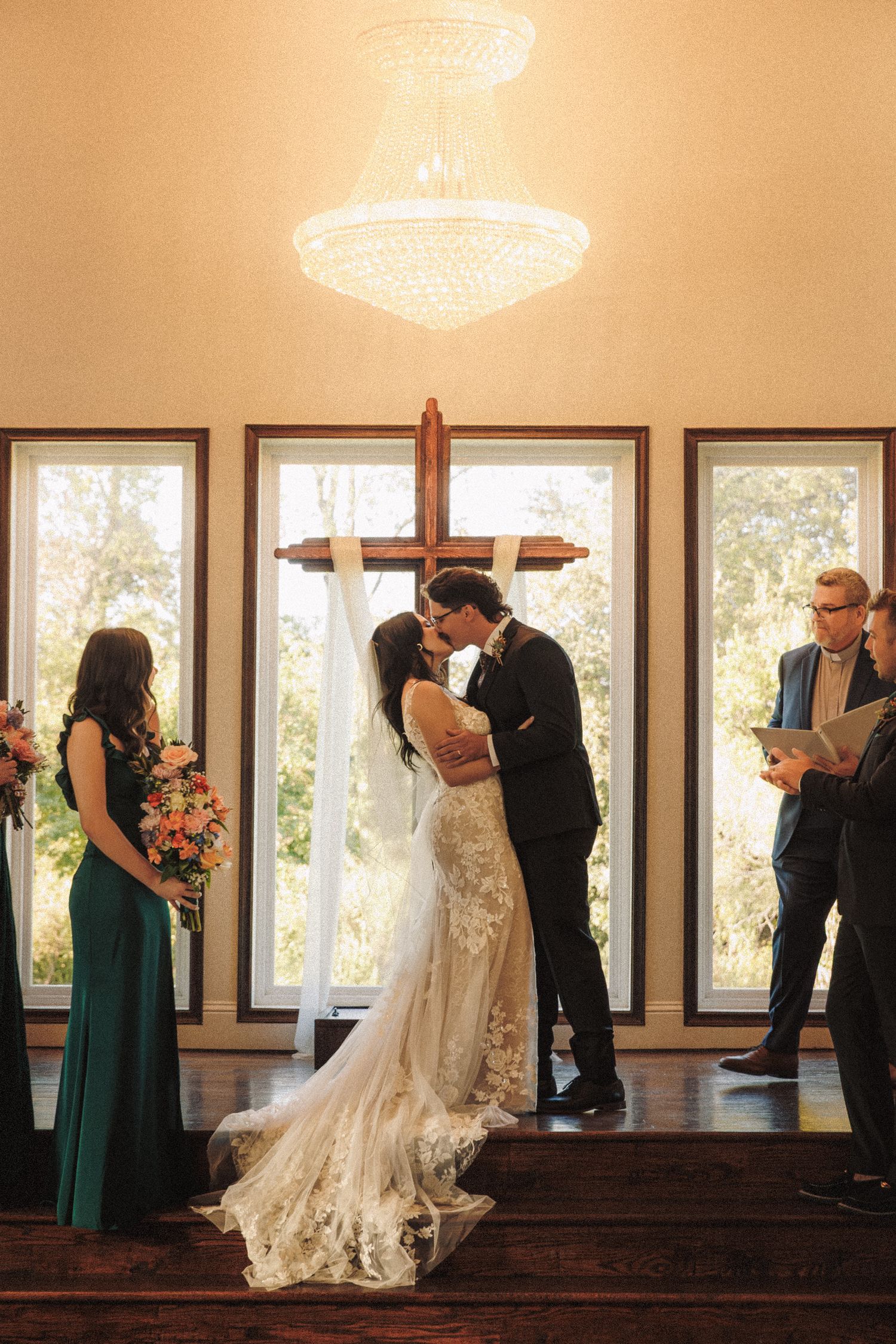 Bride and groom share their first kiss at the altar beneath a crystal chandelier and wooden cross during wedding ceremony.
