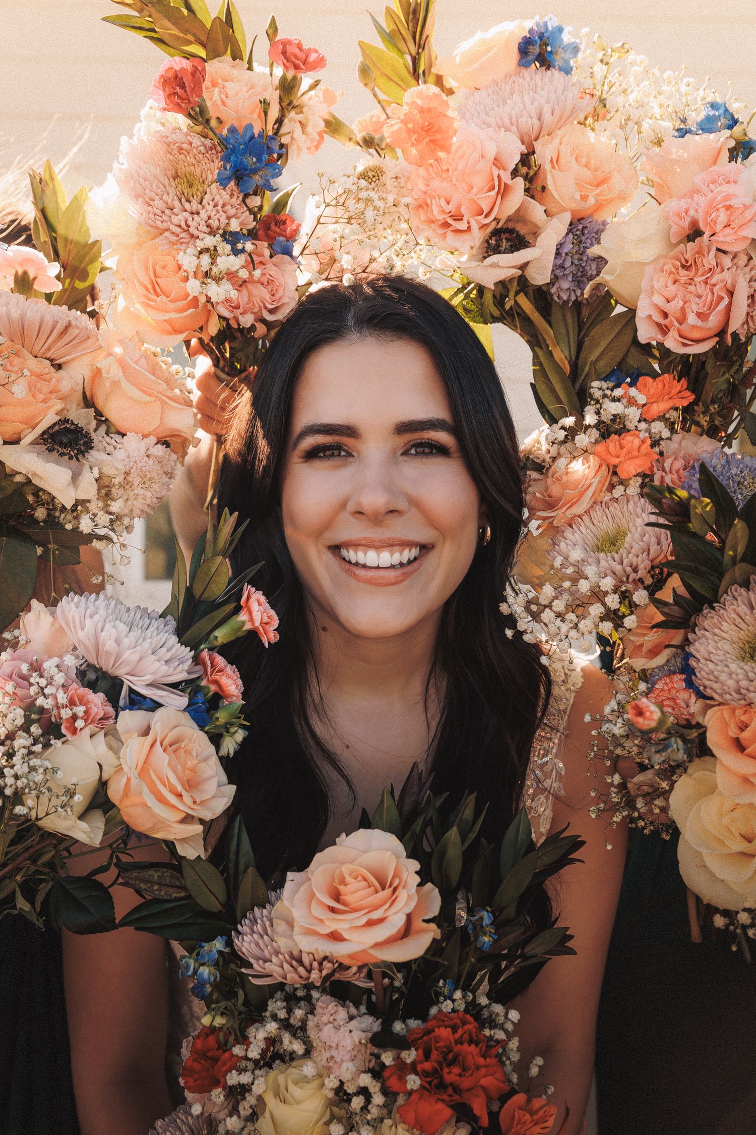 Smiling bride surrounded by colorful peach and blue floral bouquets in golden sunlight.