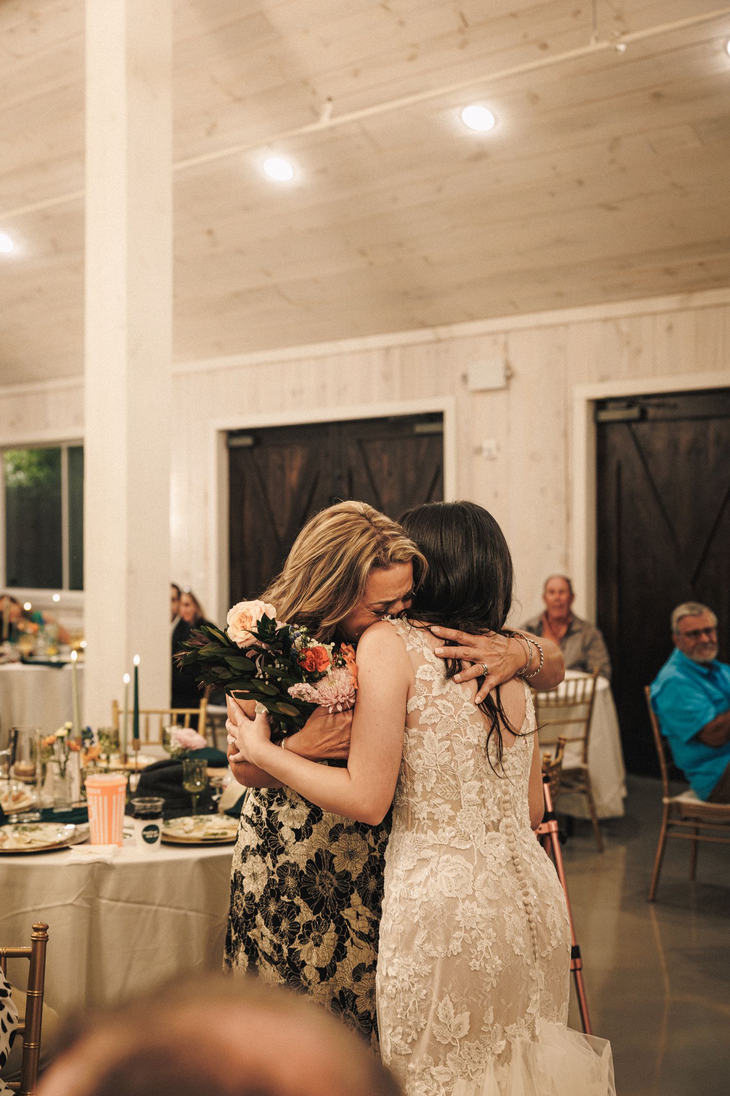 Bride in lace wedding gown shares an emotional hug with a guest in a floral dress at the wedding reception.