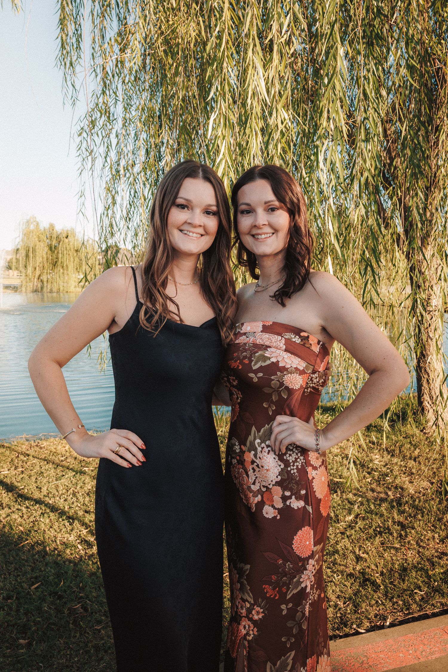 Two young women in elegant formal dresses pose together under a weeping willow tree by a lake.