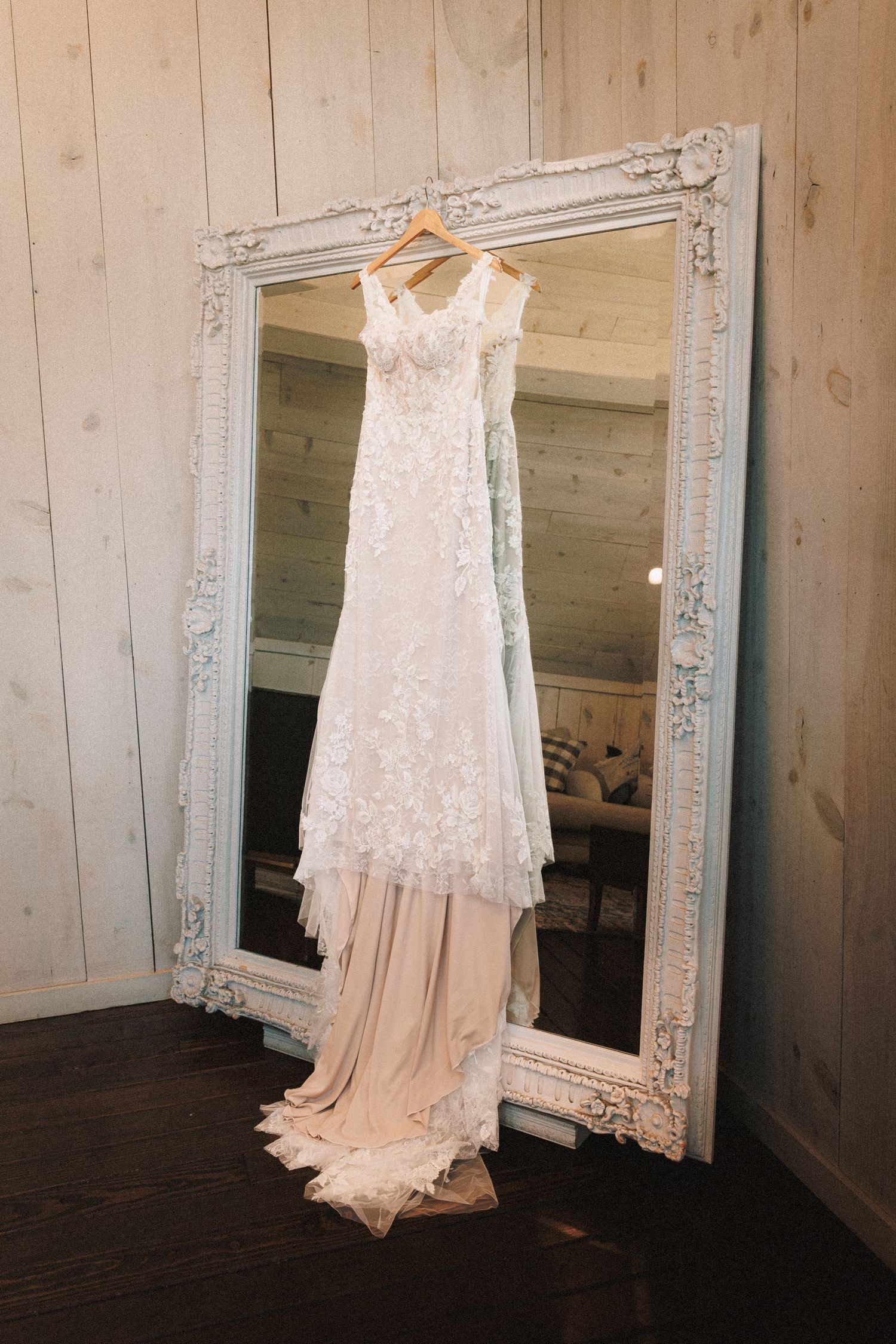 Elegant lace wedding dress hanging on an ornate white mirror frame in a rustic wood-paneled room.