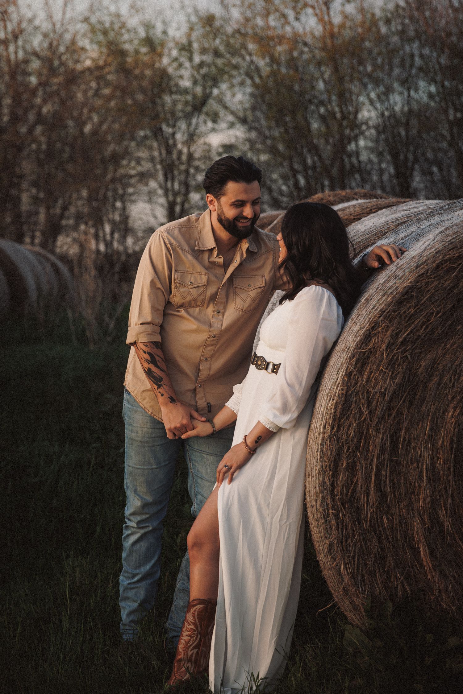 Romantic couple holding hands beside a hay bale in a rustic farm field.