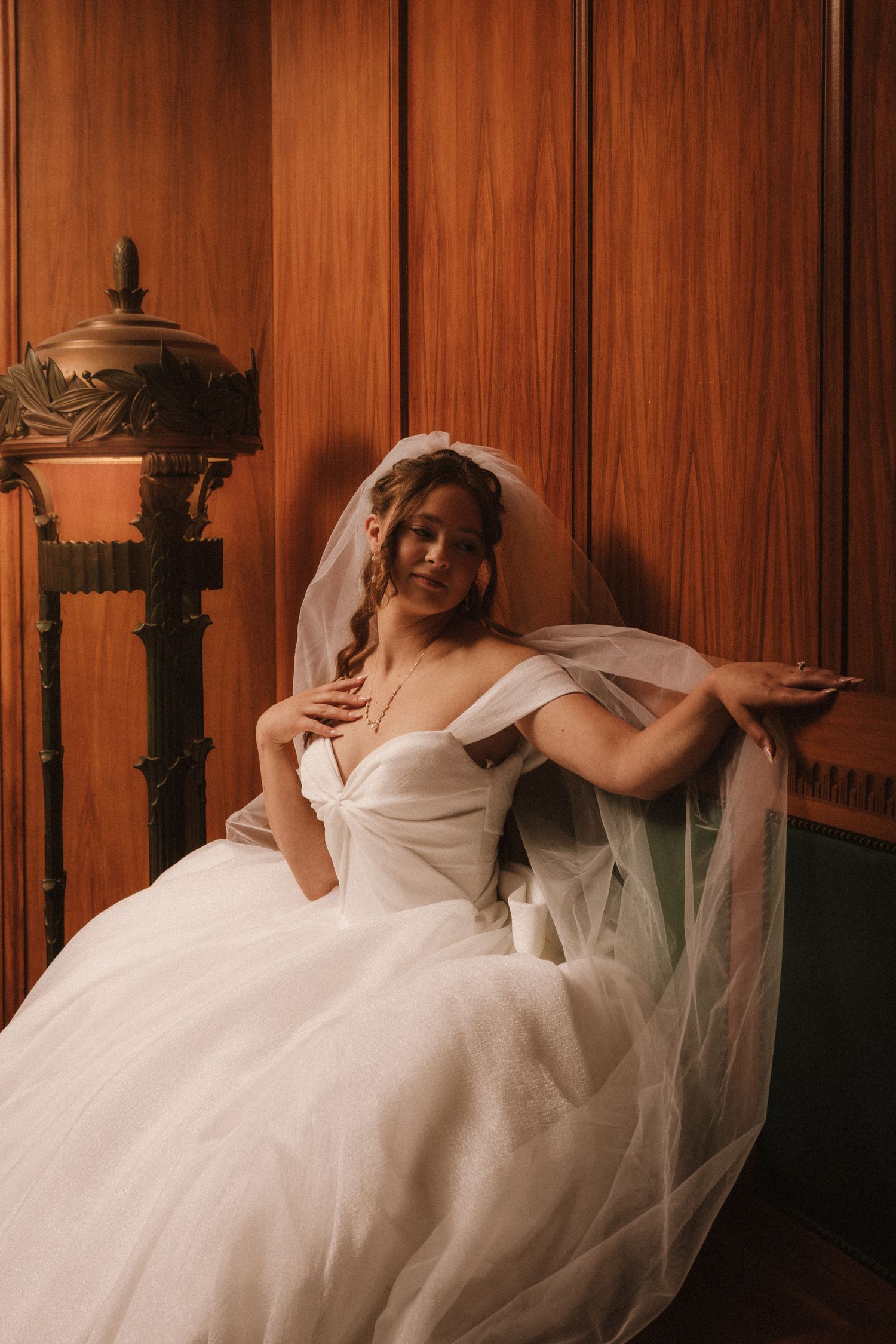 Elegant bride in off-shoulder ball gown with veil seated against warm wood-paneled wall.
