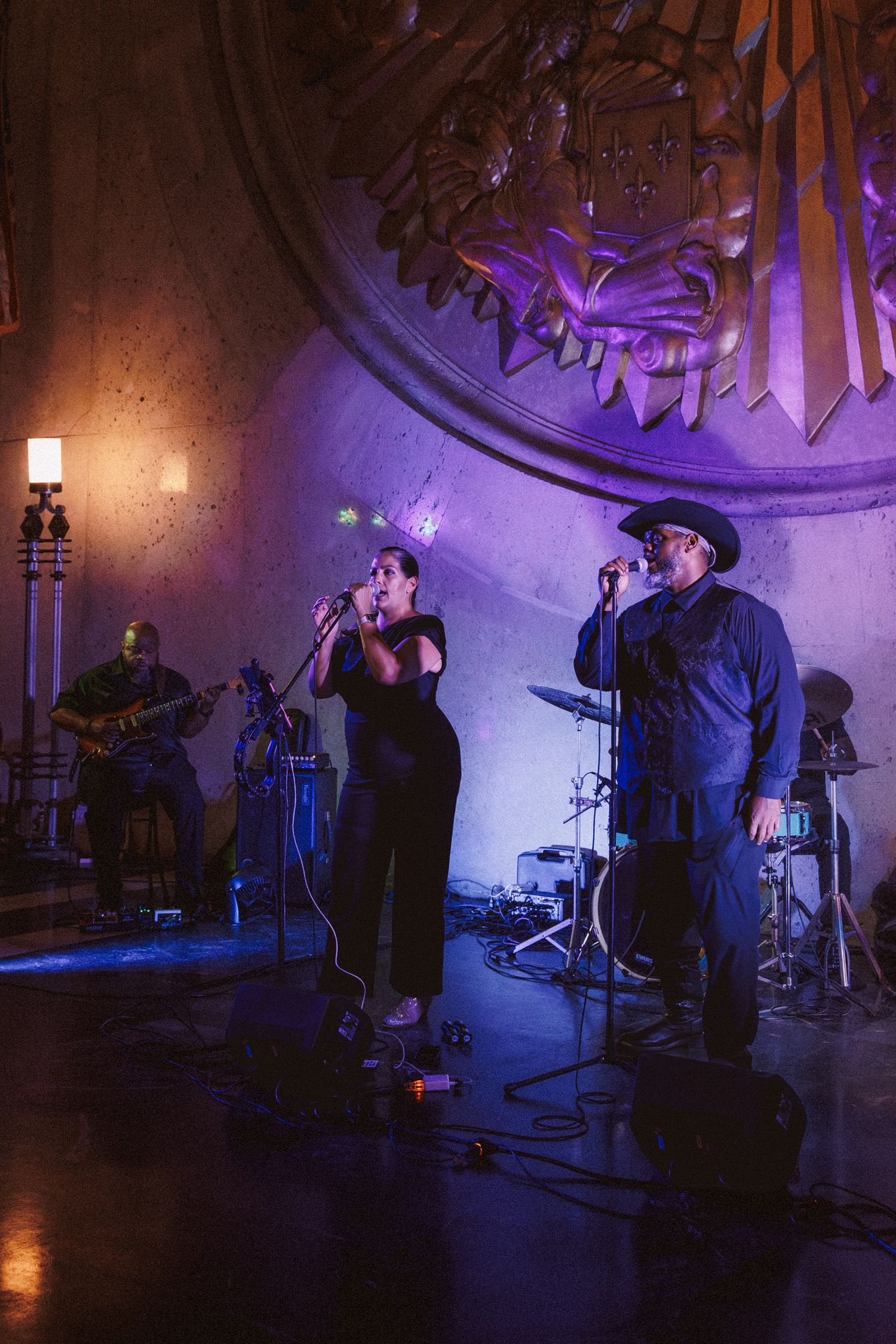 Two singers perform on a dimly lit stage with purple lighting, backed by a guitarist in an ornate venue.
