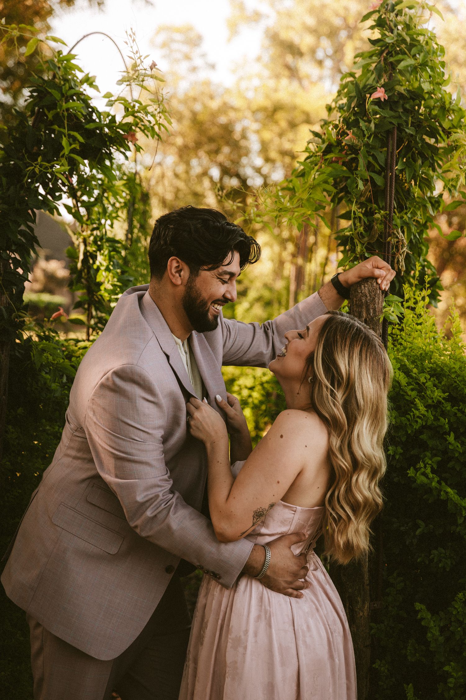 A romantic couple in beige outfits dancing playfully together in a lush garden setting at golden hour.