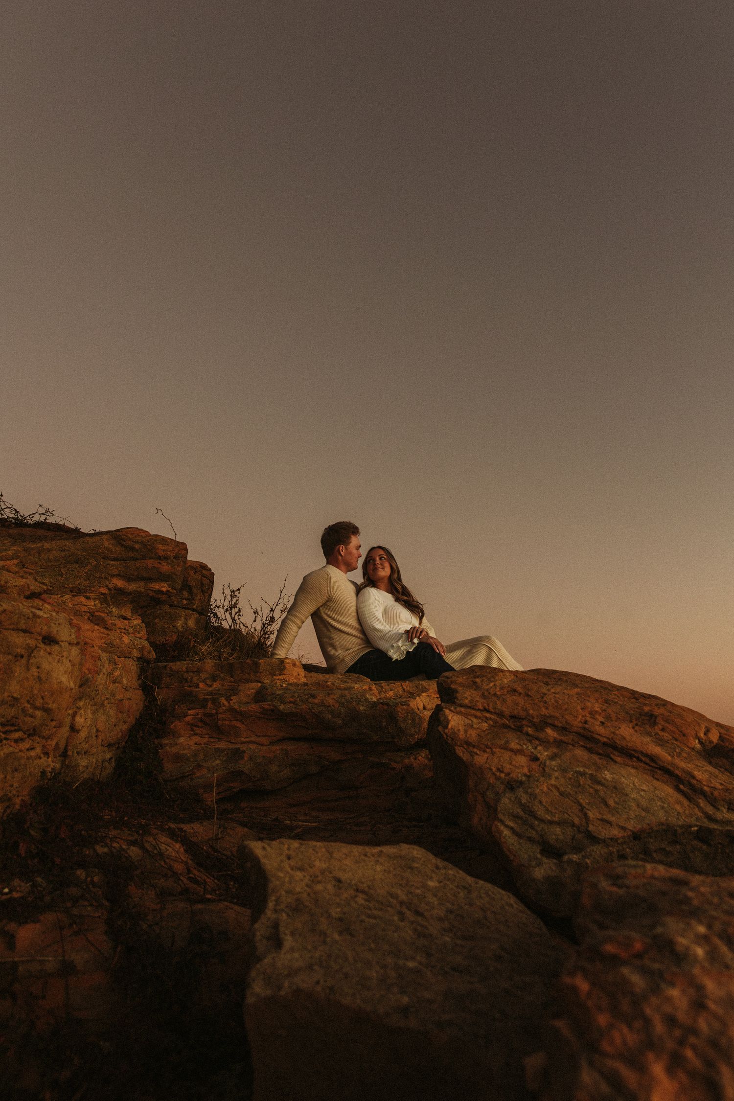 A couple sits together on a rocky cliff edge during golden hour sunset creating a romantic silhouette against the warm sky.