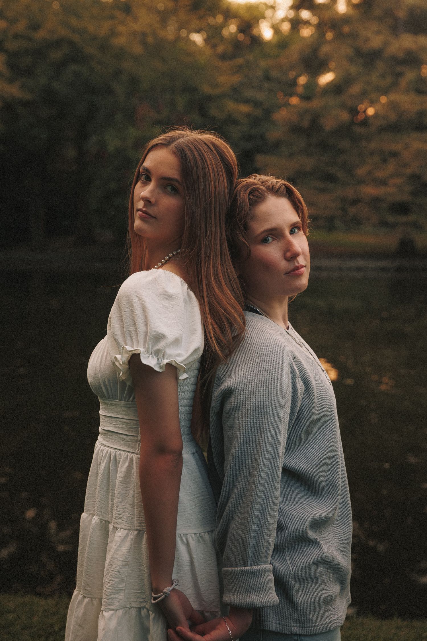 Two people in romantic poses against autumn foliage, wearing white dress and gray sweater in outdoor photoshoot.