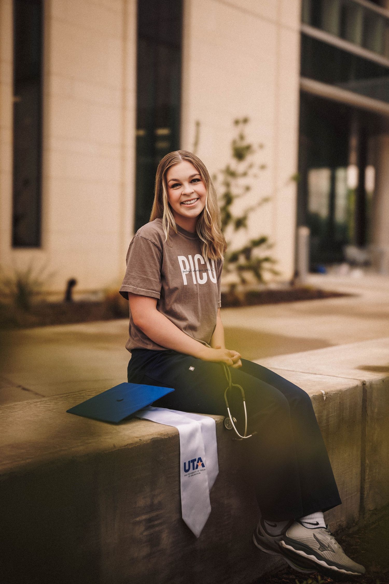 Student sits casually on concrete ledge outside modern building in brown t-shirt.