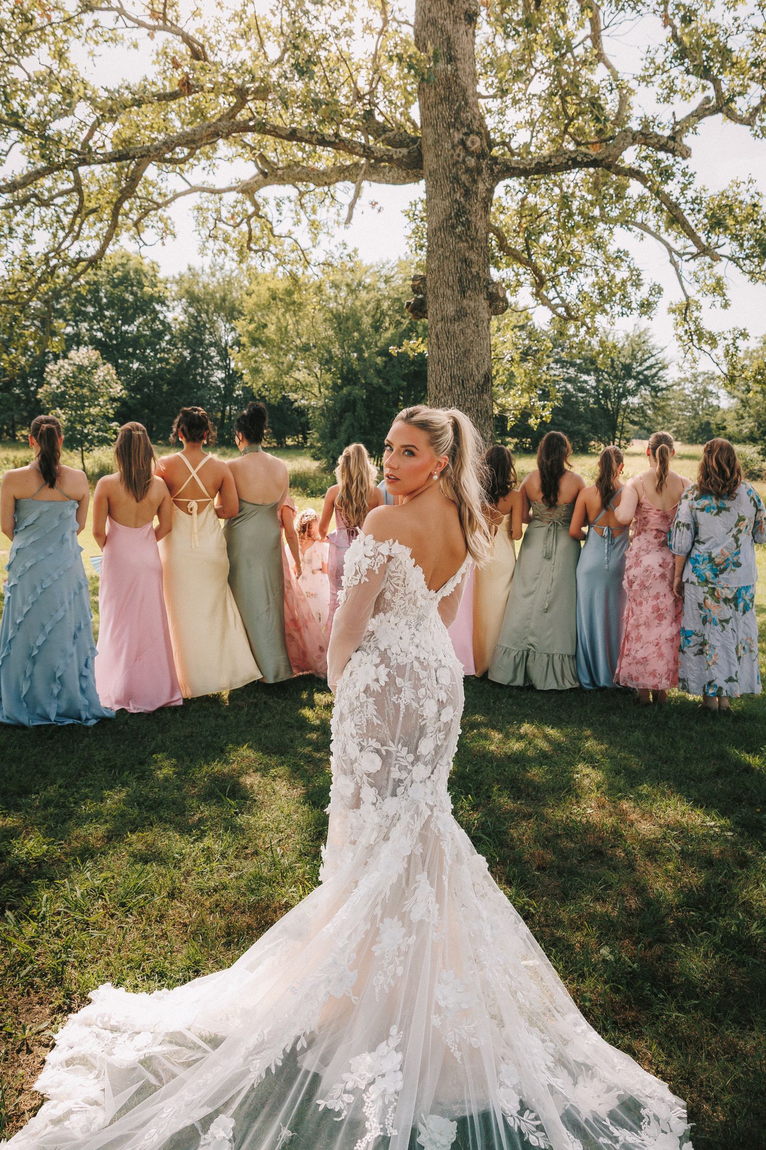 Wedding party featuring bridesmaids in pastel dresses posing outside under trees with elegant white lace bridal gown.