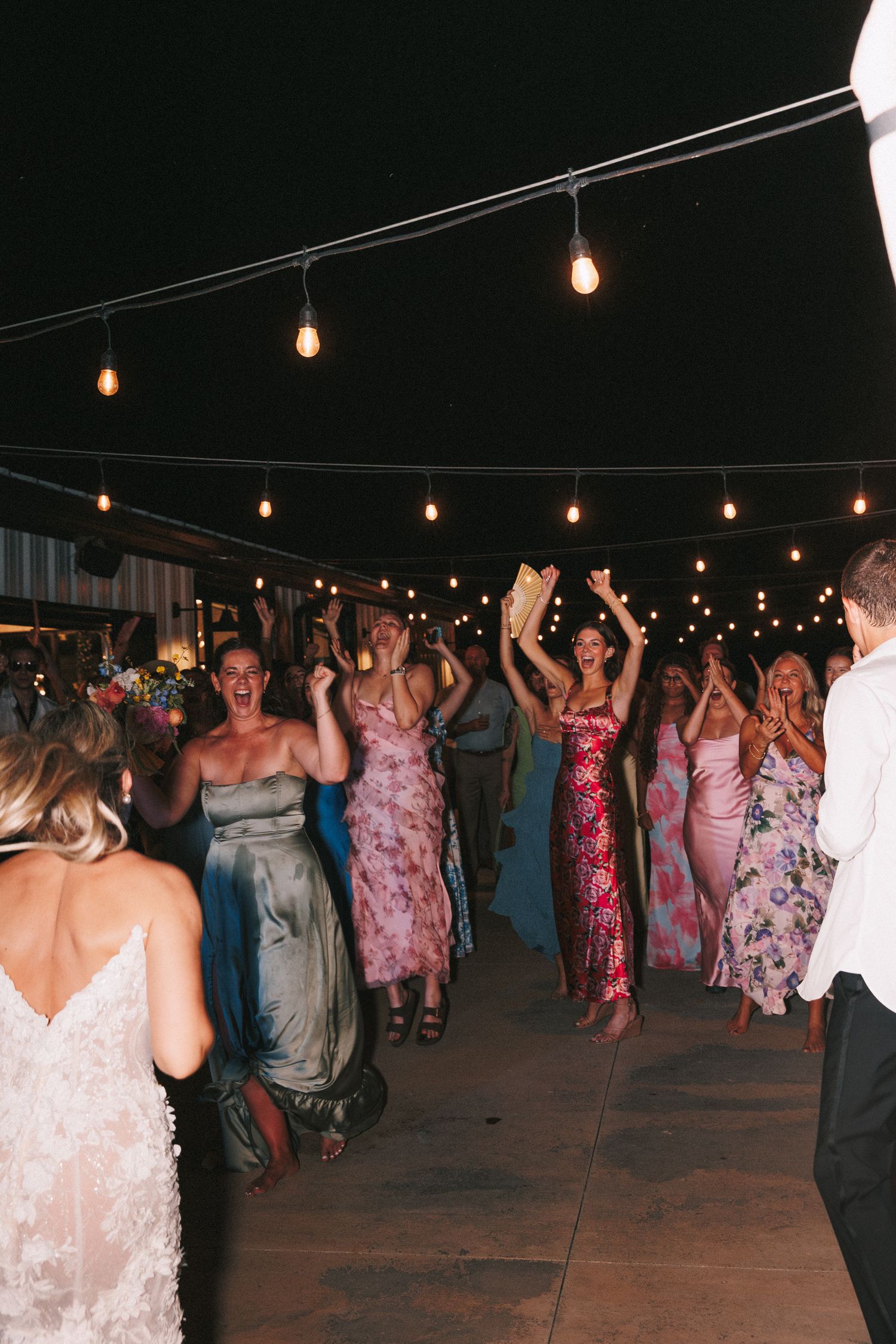 Large group dancing under string lights at an outdoor wedding reception with guests wearing colorful dresses.