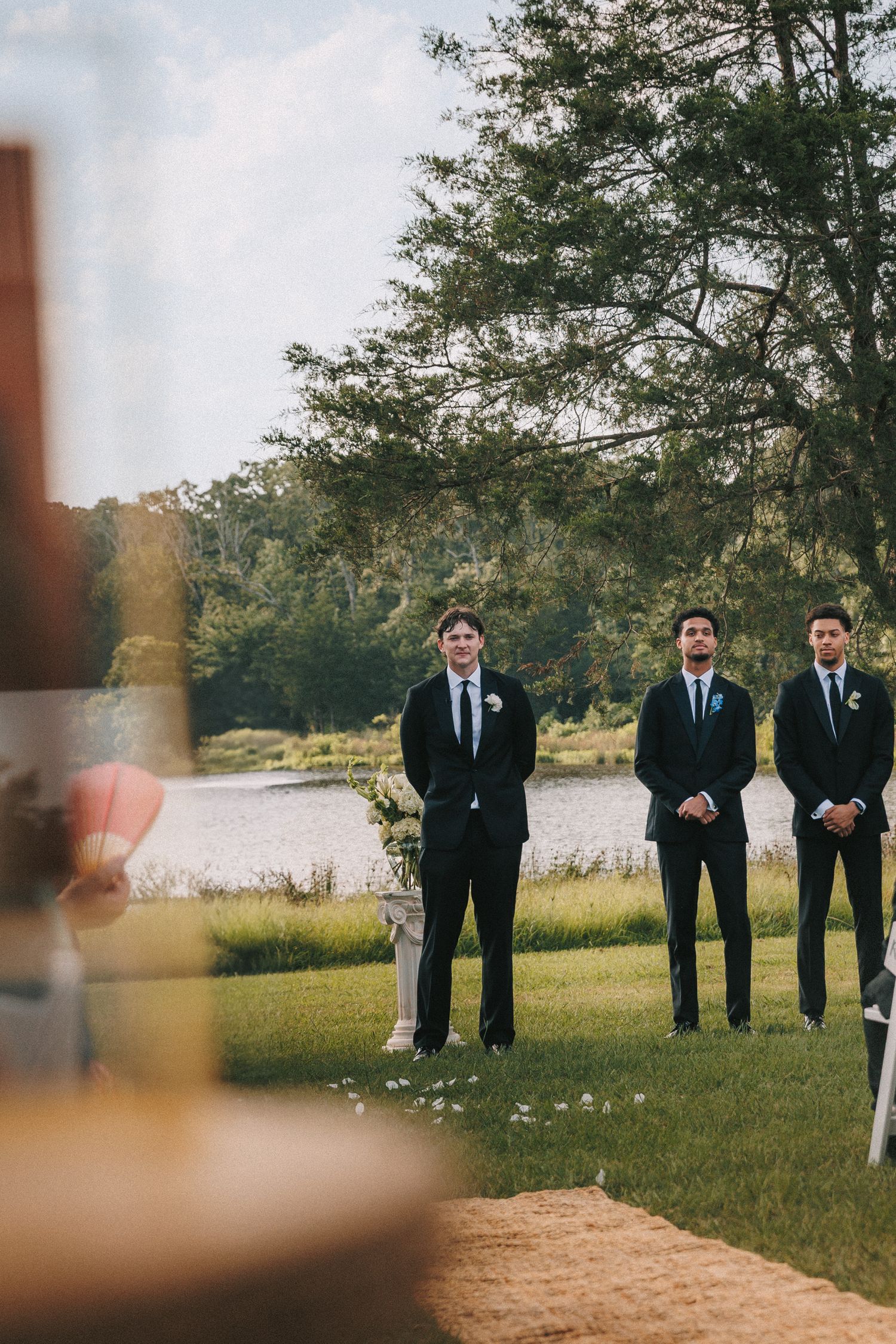 Groomsmen in dark suits stand outdoors against a scenic lakeside landscape.
