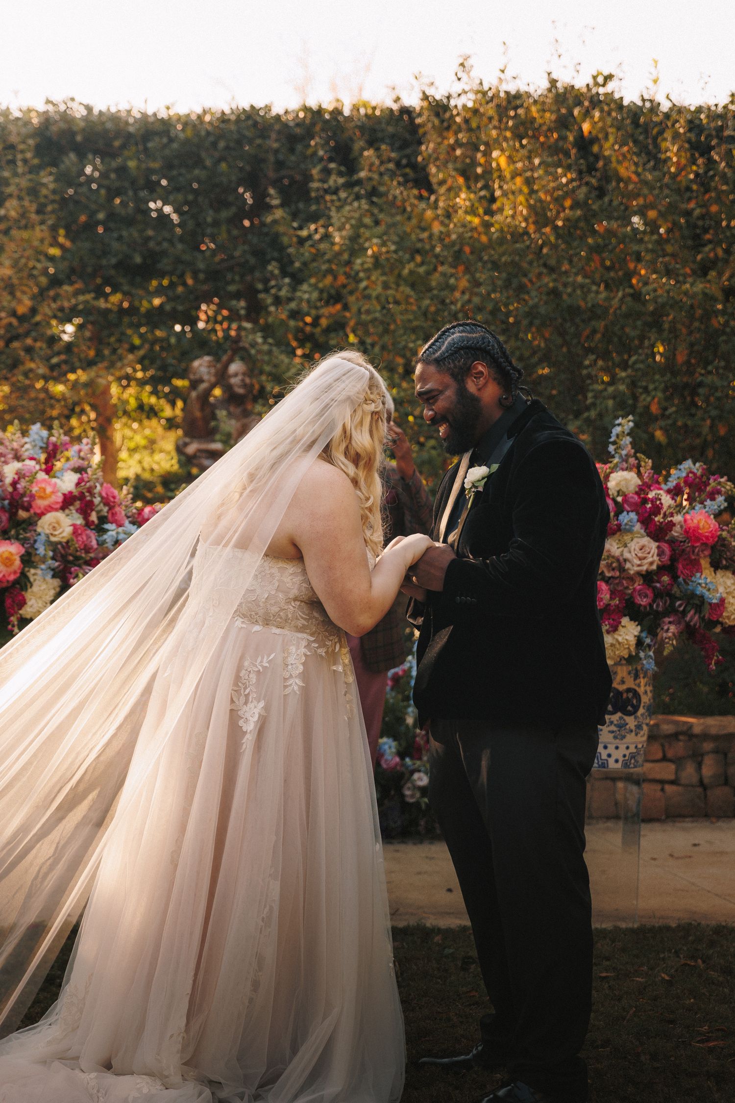 Bride in lace veil and gown exchanges rings with groom amid colorful floral arrangements outdoors.