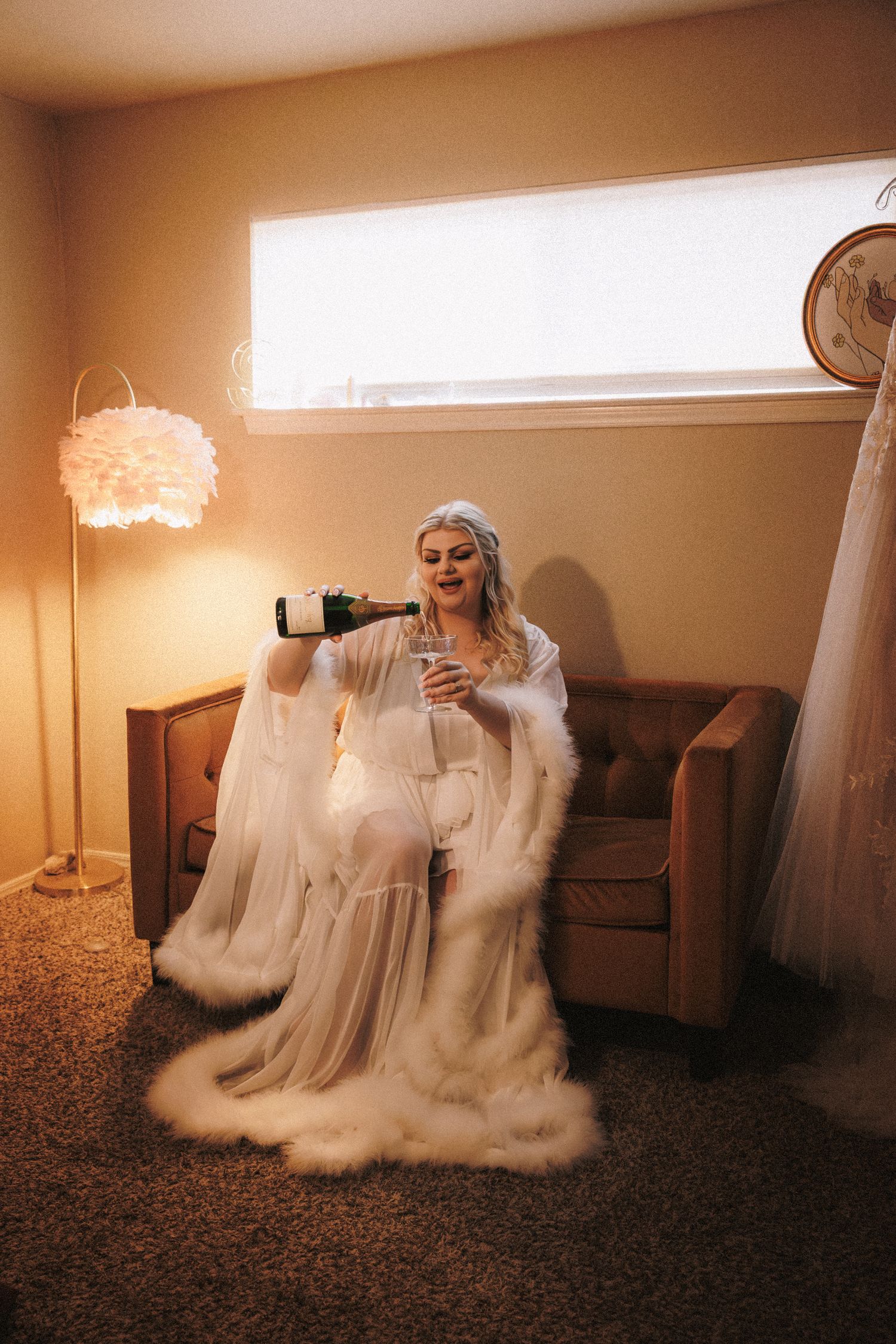Bride in sheer white robe and fur trim pouring champagne while seated in cozy vintage room.