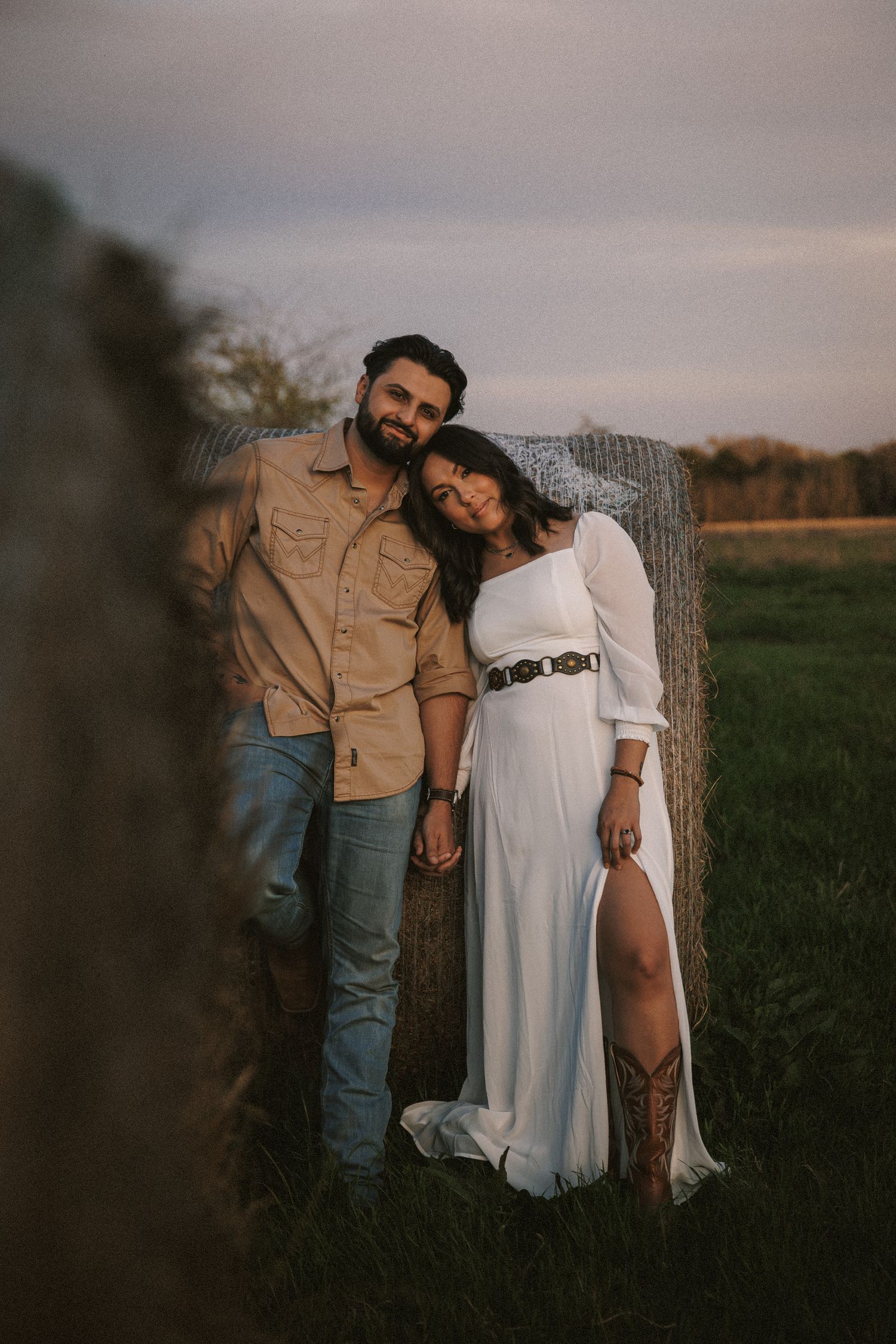 Couple posing together in a field leaning against a hay bale at sunset, woman in white dress, man in tan shirt.