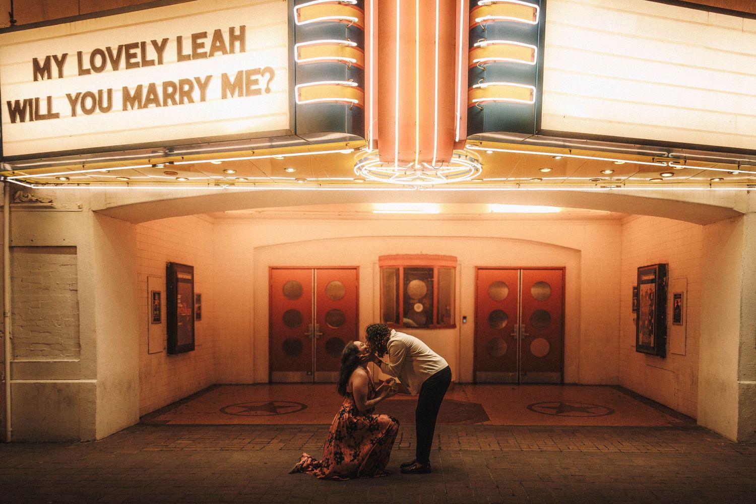 Couple embracing under a bright theater marquee displaying My Lovely Leah Will You Marry Me at night.
