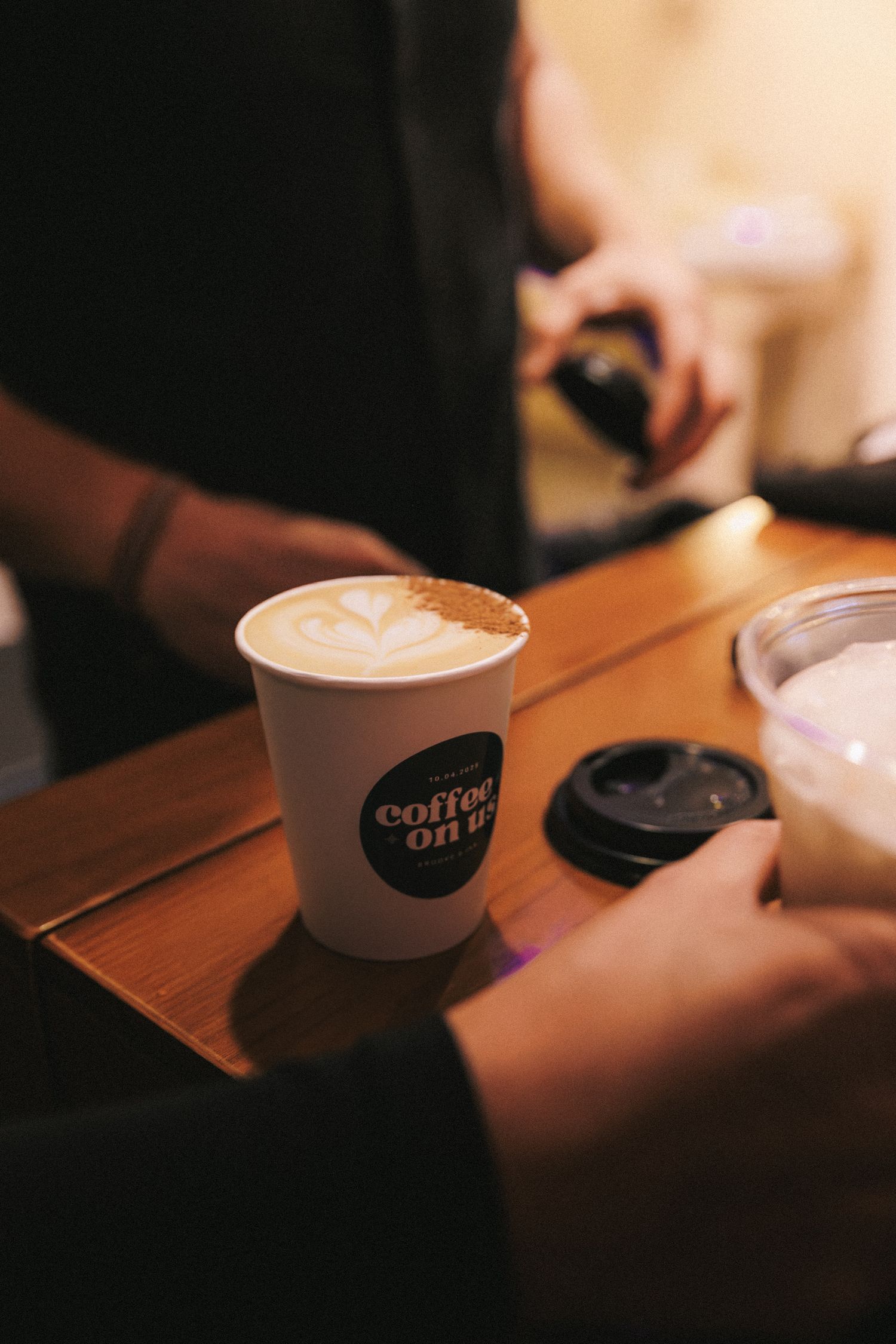 Barista serving a latte art coffee in a branded takeaway cup on a wooden counter.