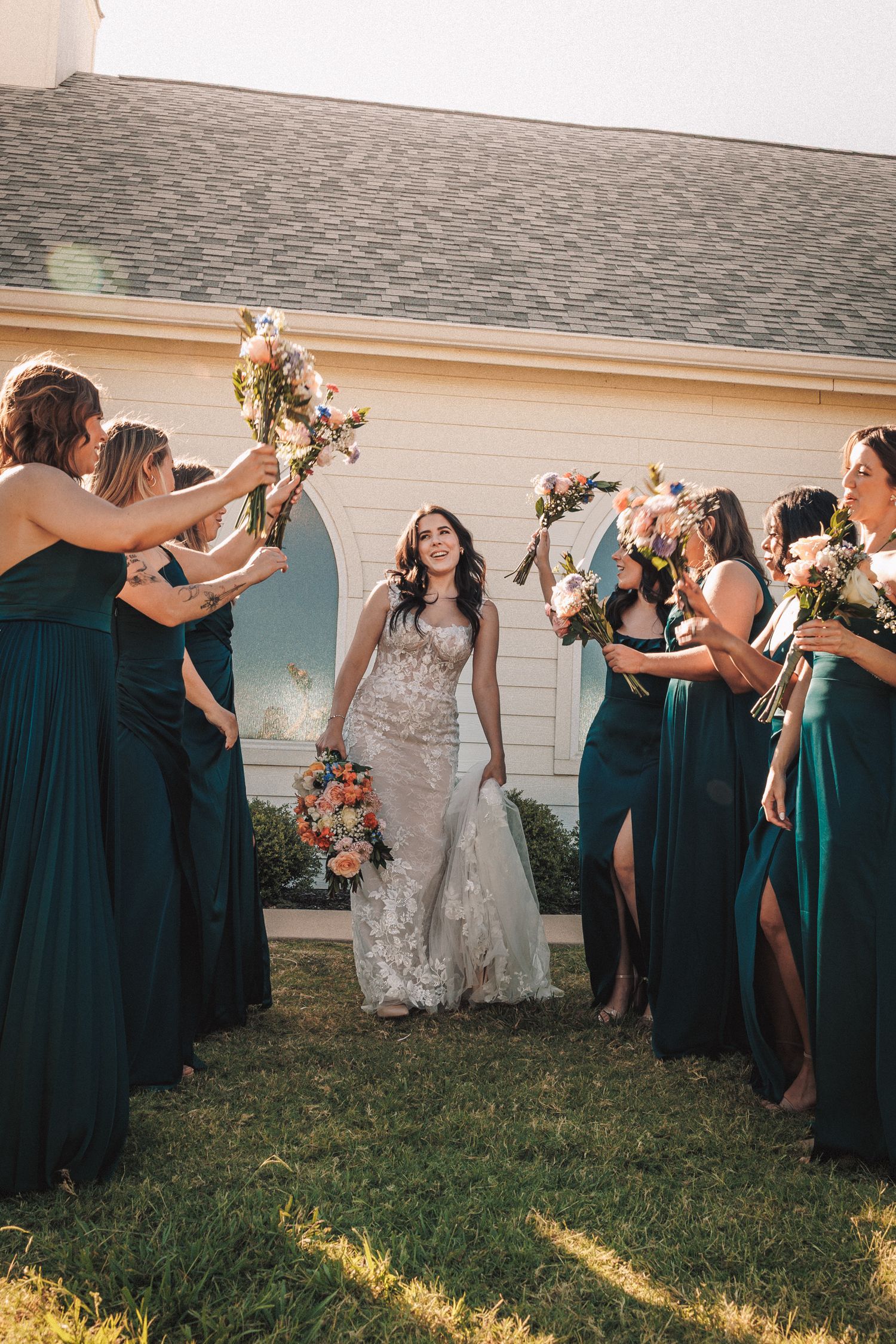 Bride walks between bridesmaids in emerald green gowns raising colorful bouquets outside a white chapel.