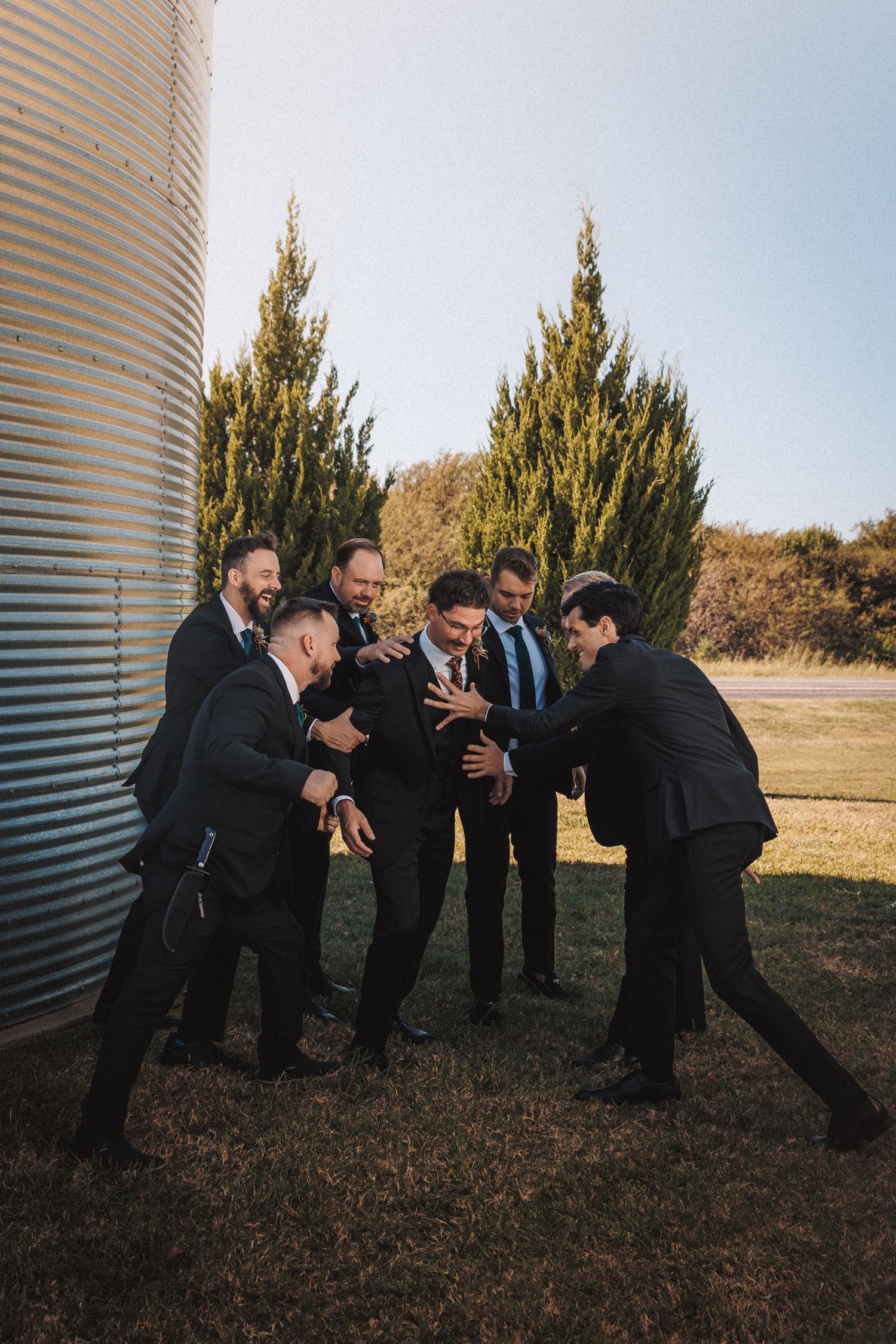 Groomsmen in dark suits playfully surrounding the groom outdoors near a grain silo.
