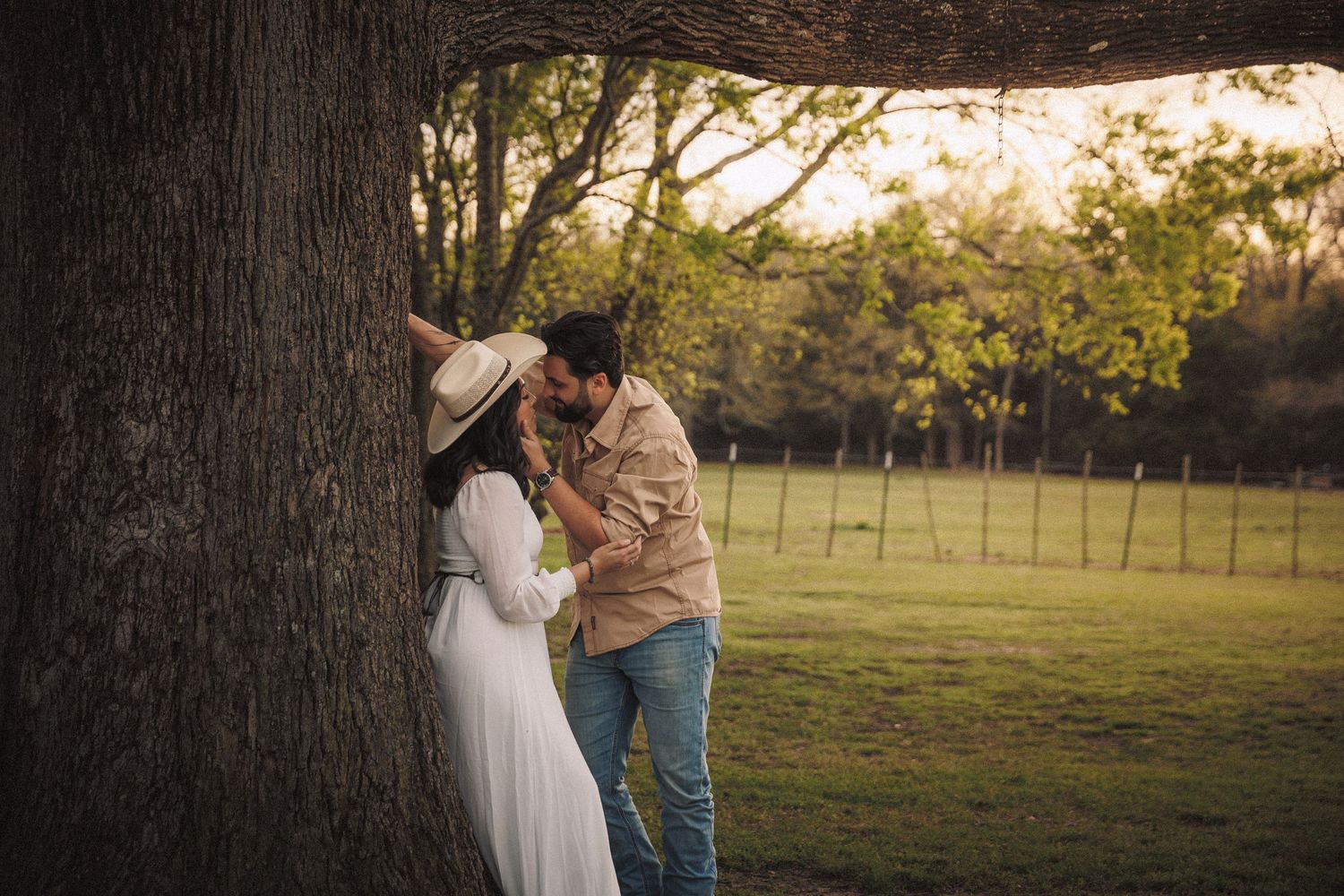 Couple sharing a romantic moment beneath a large tree in a sunlit green field.