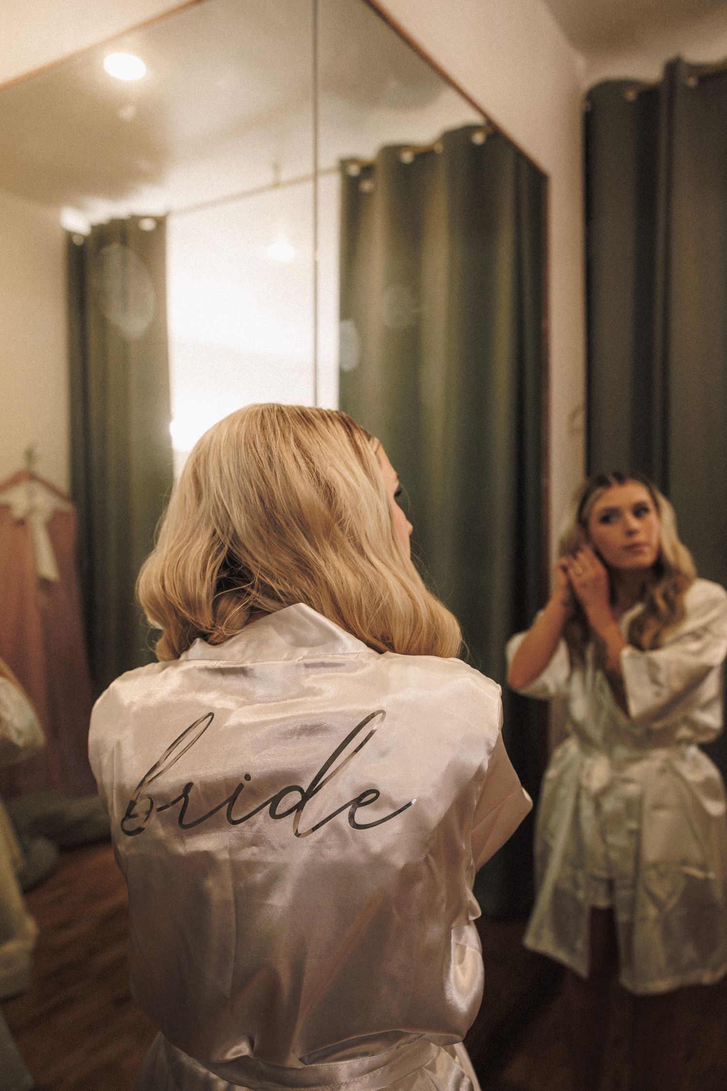 Blonde bride in white satin robe with script bride text reflected in mirror during wedding preparations.