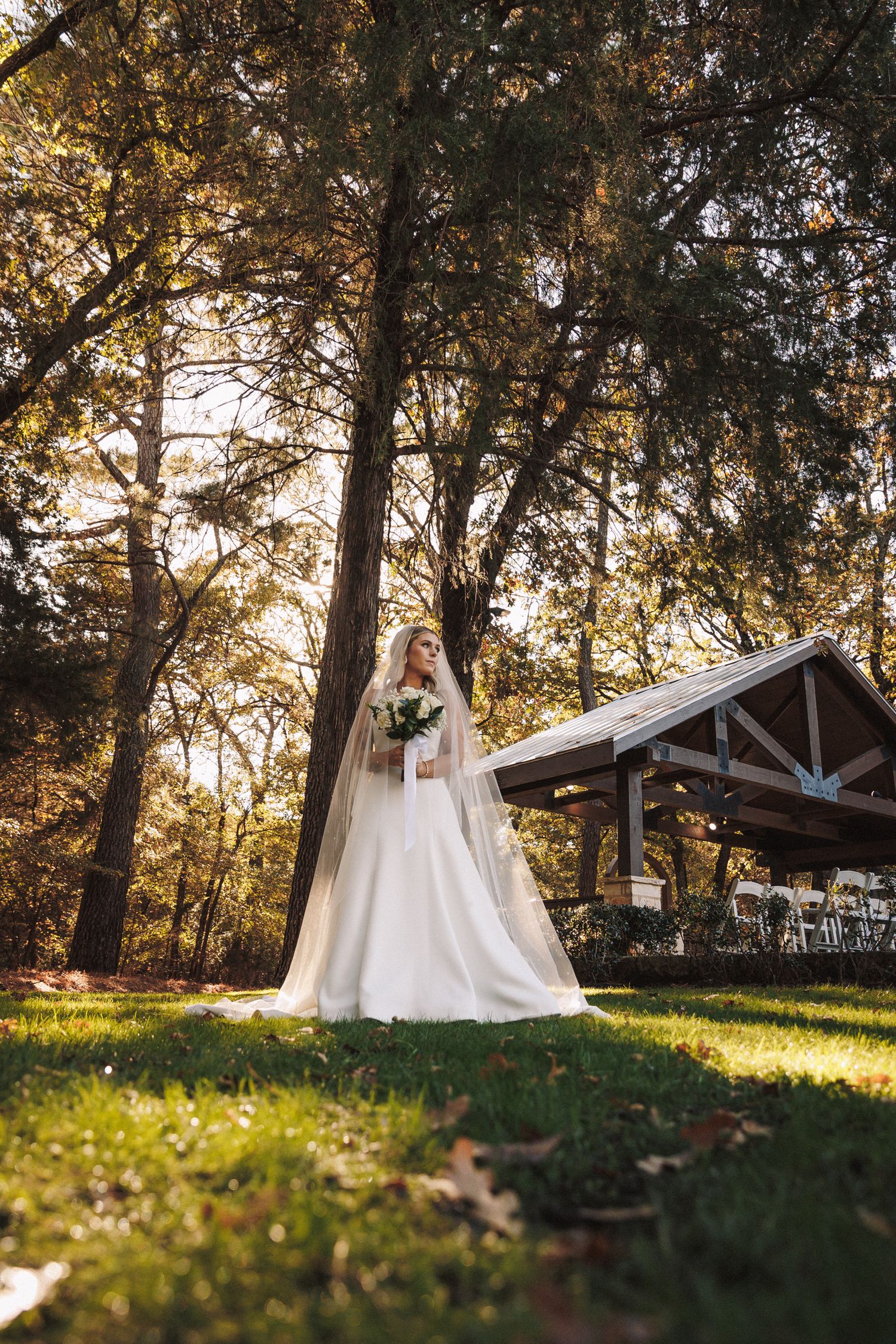 Elegant bride in white gown and veil holding bouquet among tall trees at outdoor venue.