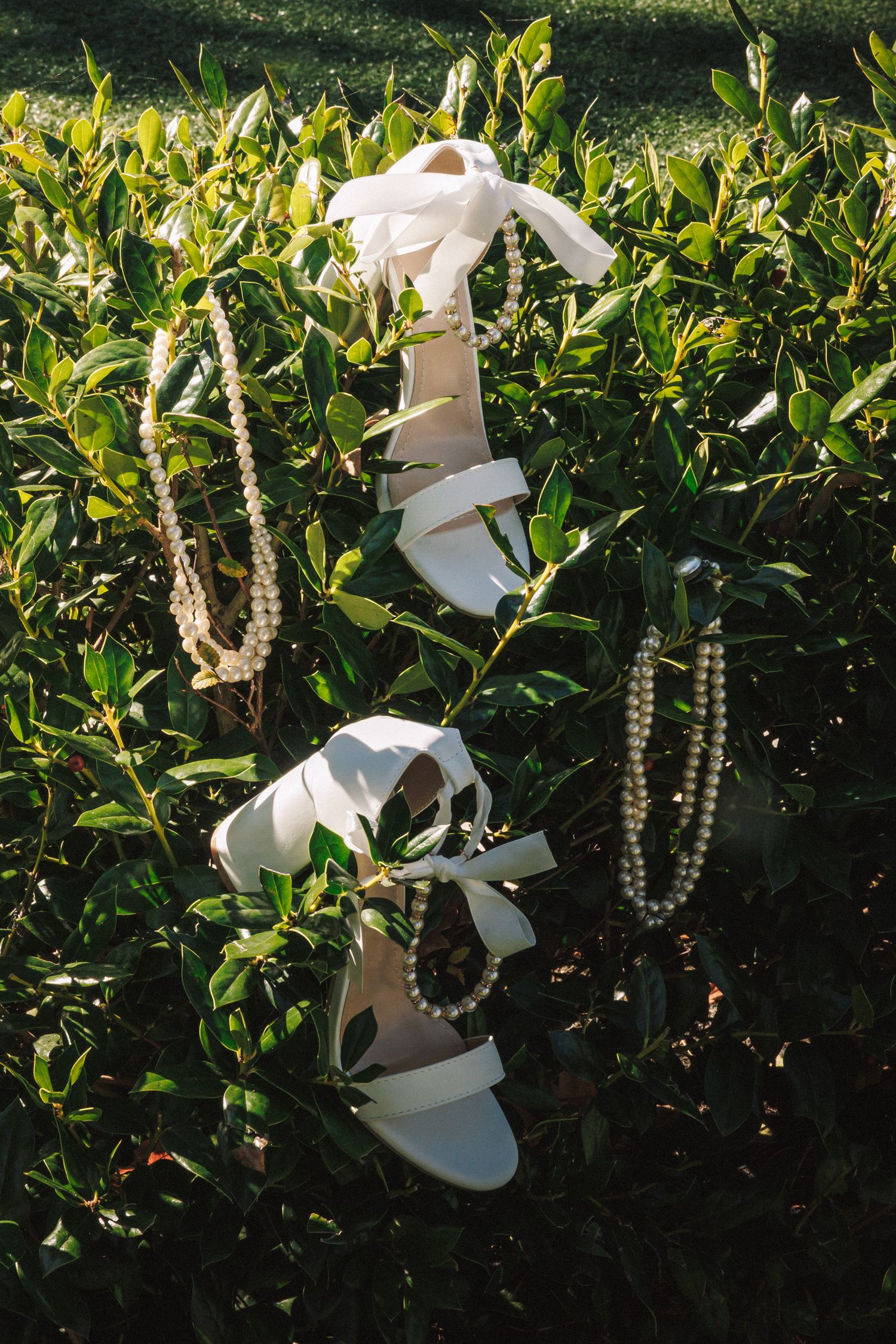 White bridal heeled sandals with ribbon ties and pearl jewelry displayed on lush green bushes outdoors.