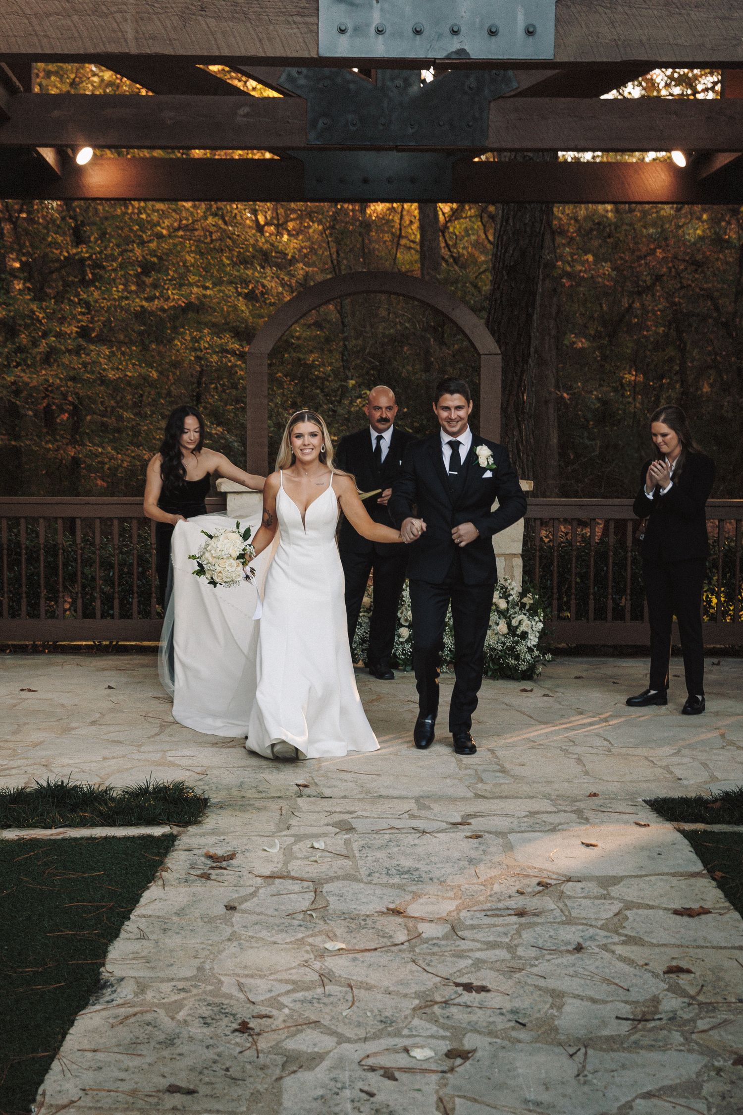 Newlyweds joyfully walk back down aisle after wedding ceremony under rustic wooden arch at dusk.