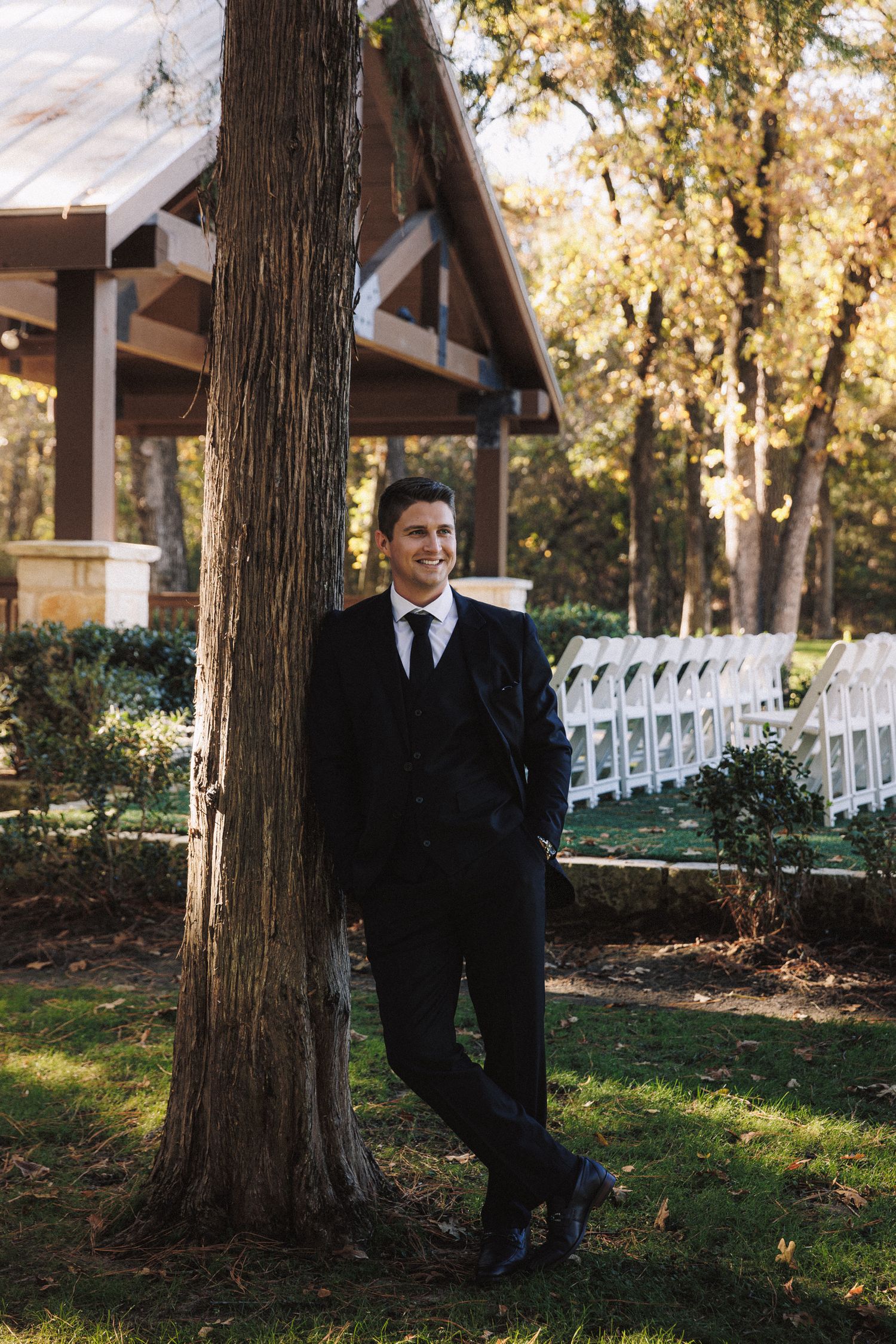 Groom in black suit leaning against tree near outdoor wedding ceremony chairs in autumn setting.