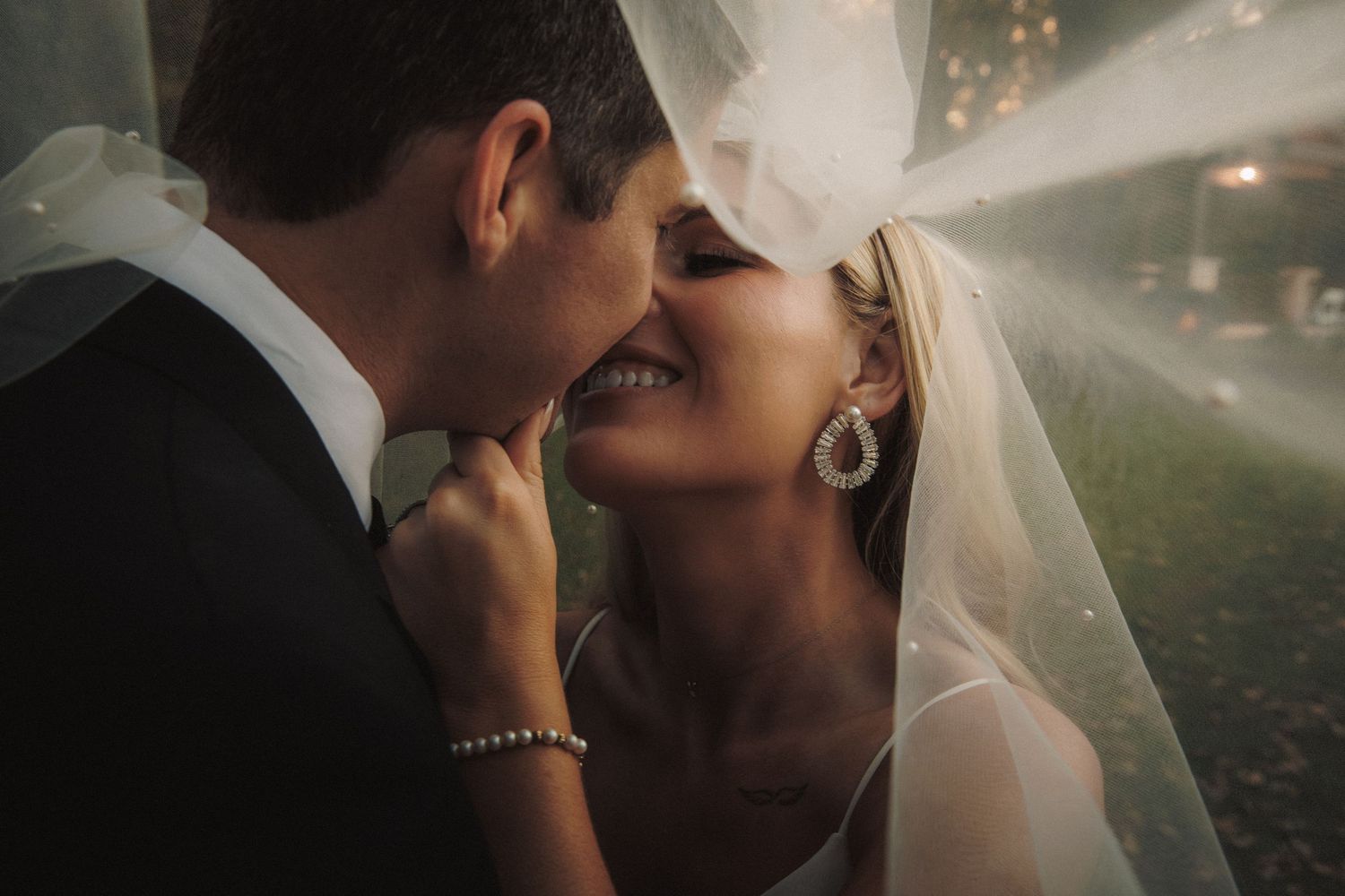 Bride smiling under veil as groom leans in for a kiss in romantic portrait.