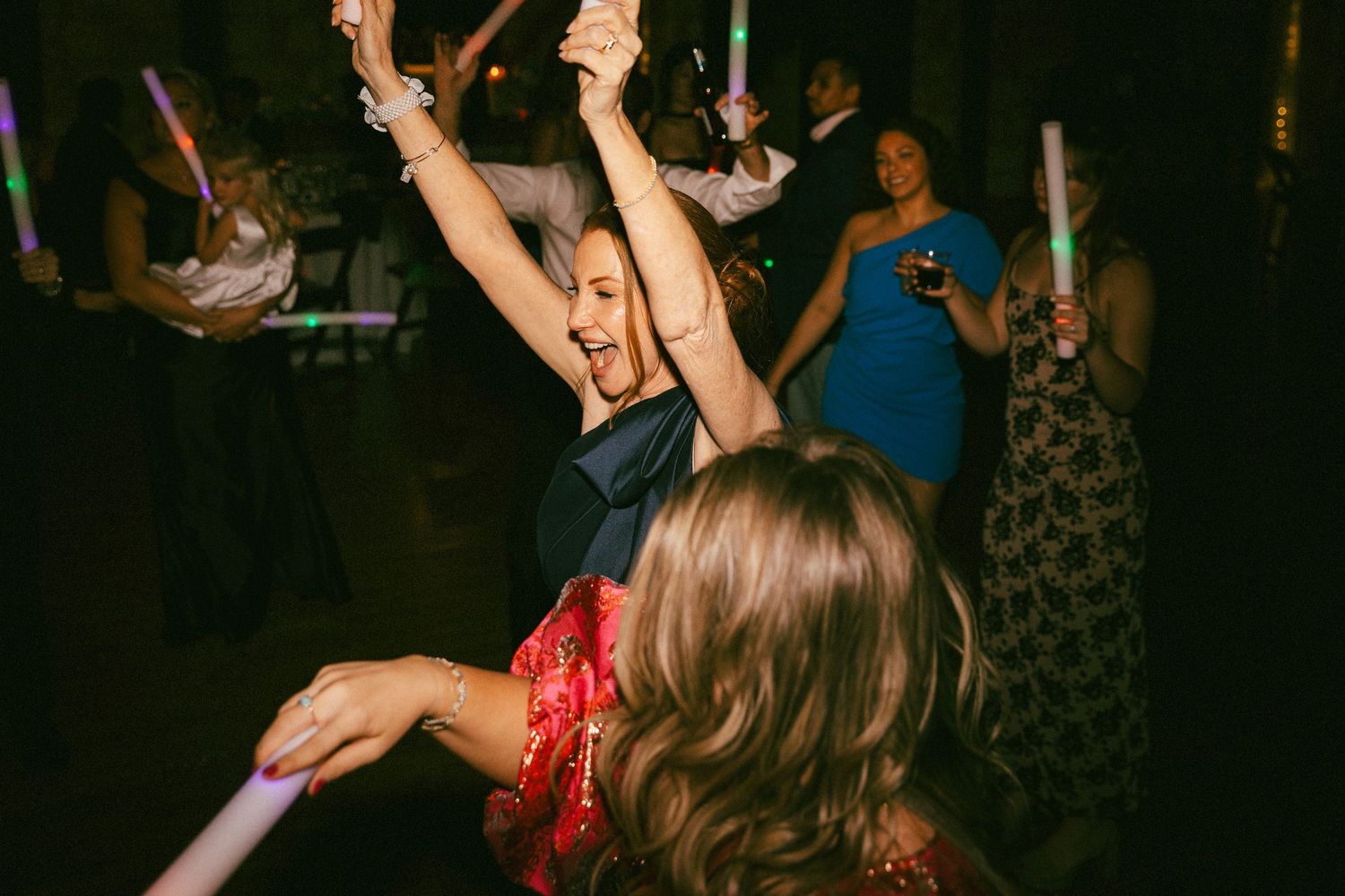 Excited wedding guest in navy dress dancing with arms raised at lively indoor wedding reception.