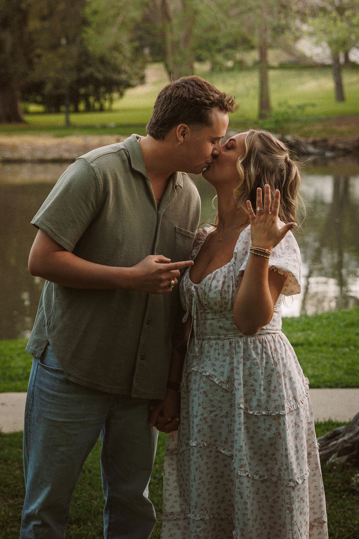 Couple kissing and showing engagement ring by a serene pond in a park.