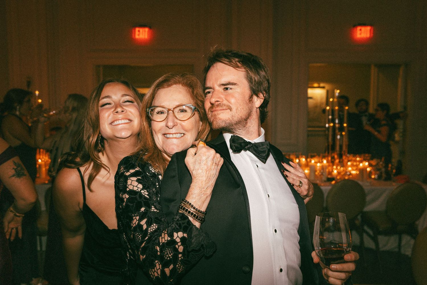 Three guests laughing and embracing together at a warm candlelit wedding reception.