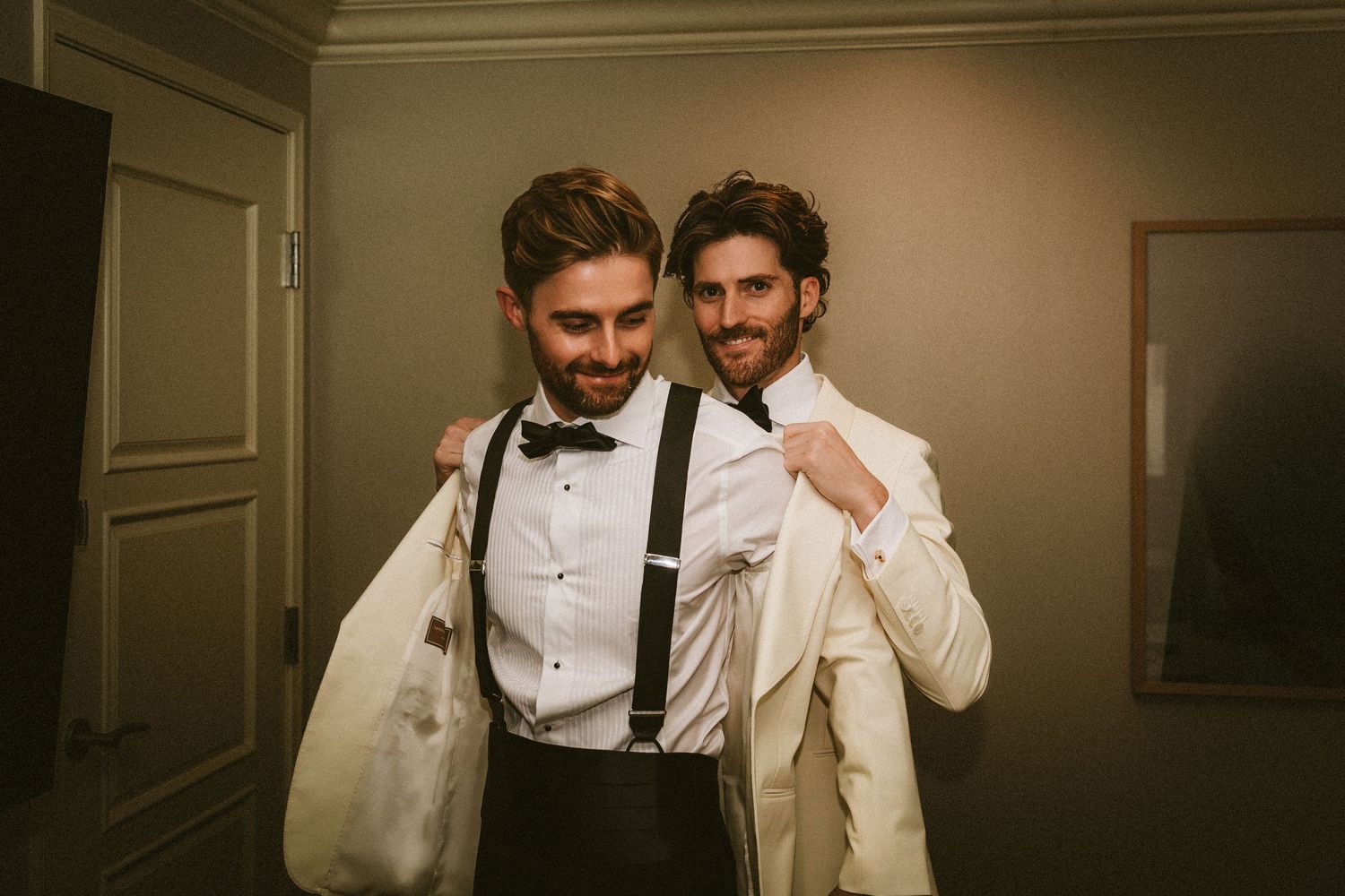Two grooms in white tuxedos with bow ties and suspenders smiling together before their wedding.