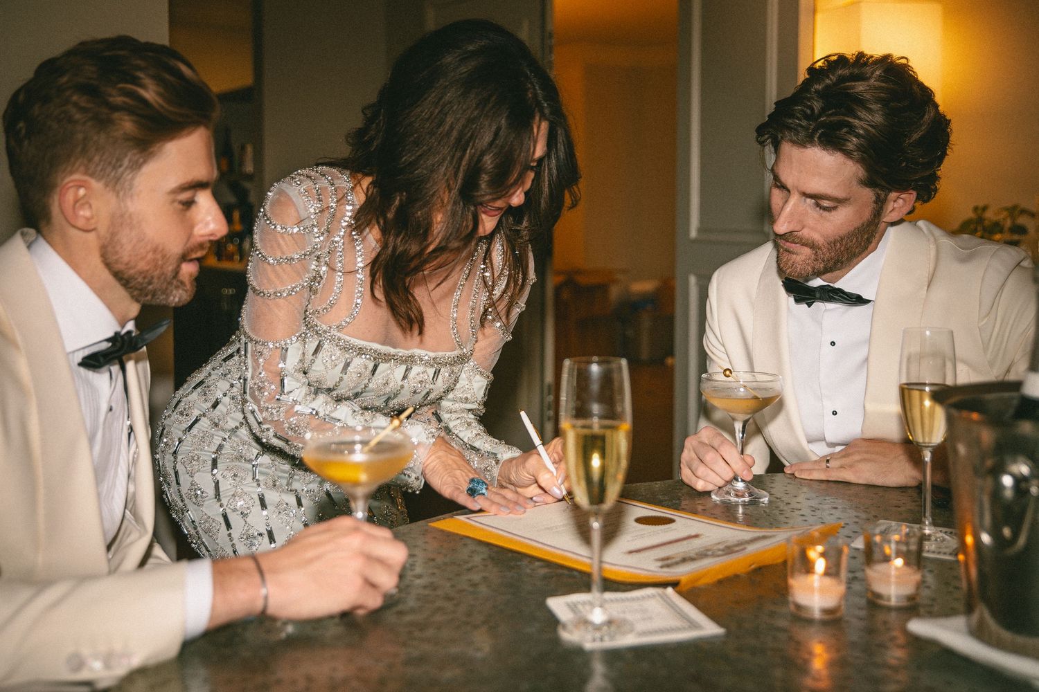 Two grooms in ivory tuxedos sign wedding documents with female guest at candlelit bar holding cocktails.