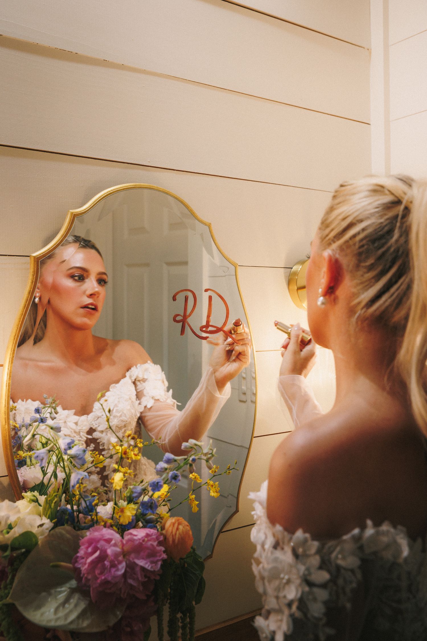 Bride getting ready at vanity mirror with colorful floral arrangements in foreground.
