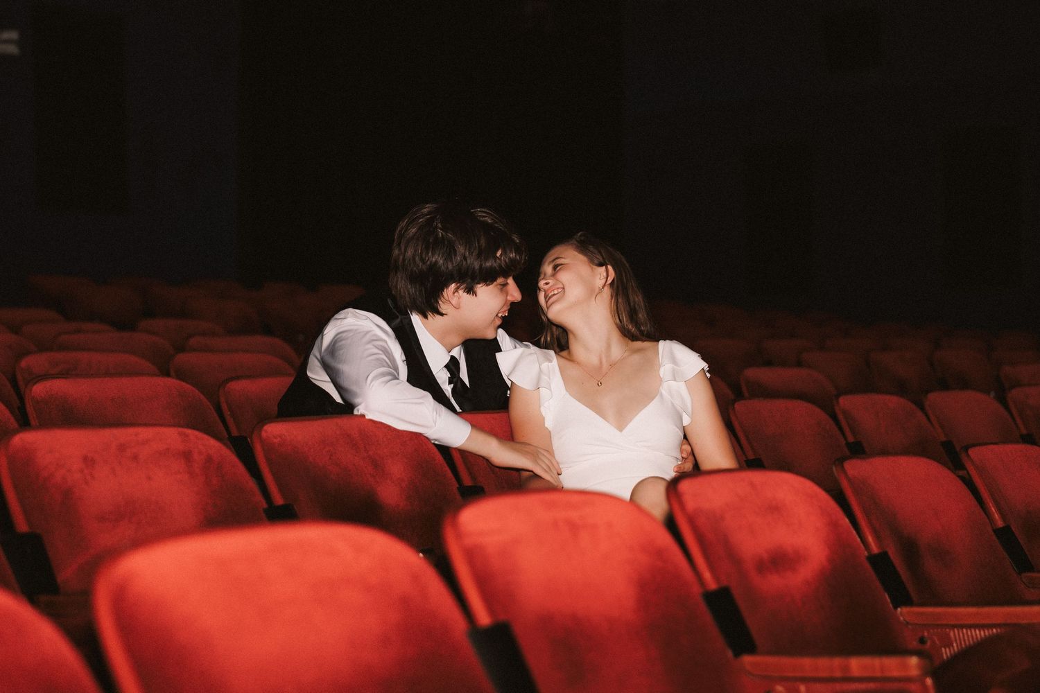 A couple sharing an intimate moment together in the empty red seats of a dimly lit theater.