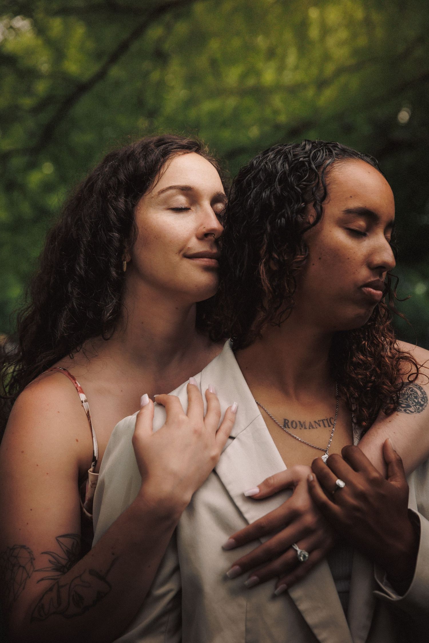 Two people with curly hair in light colored tops share an intimate embrace against a blurred forest background.