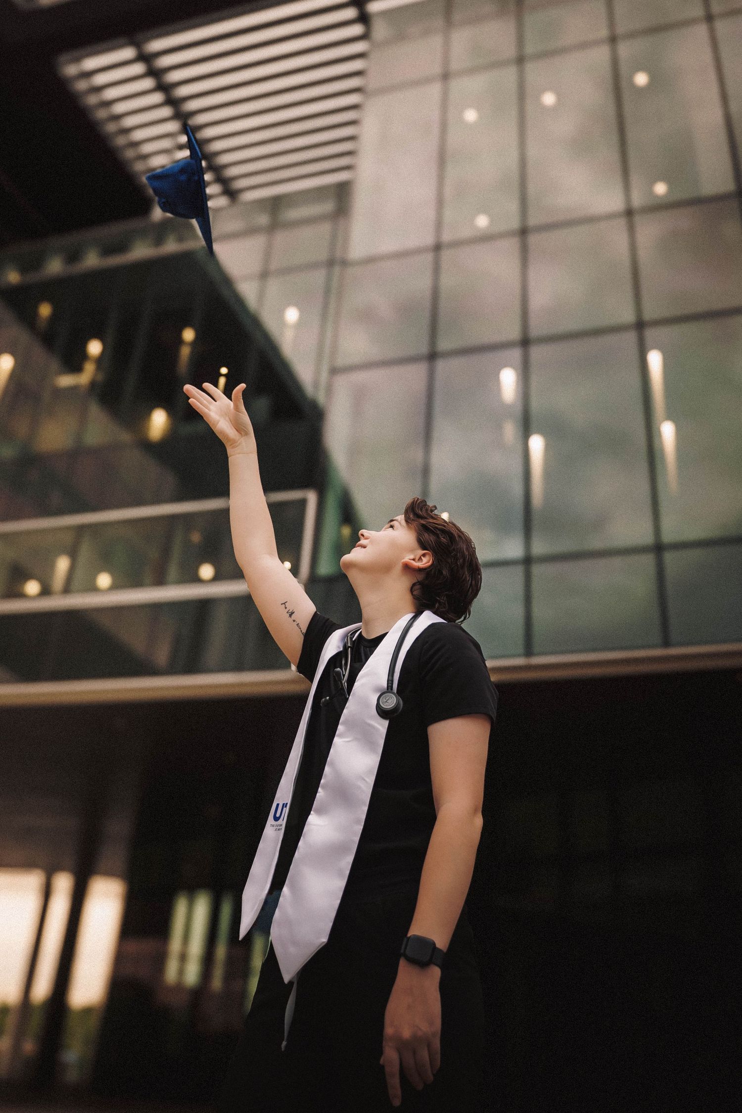 Graduate celebrates achievement outside modern building at dusk with dramatic upward pose.