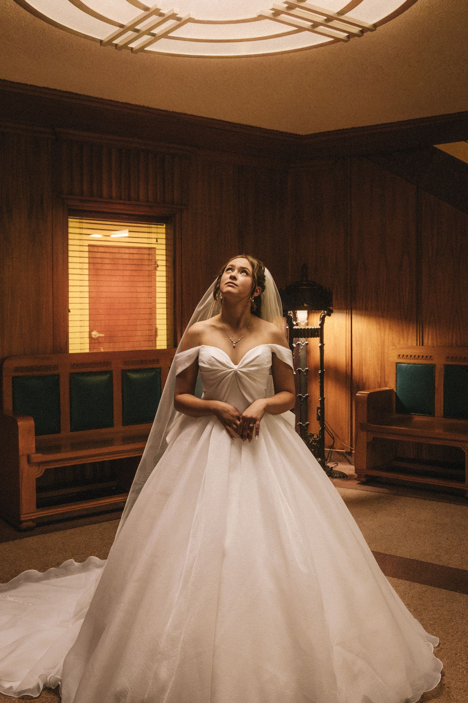 Bride in off-shoulder ballgown and veil stands in warmly lit wood-paneled room, gazing upward elegantly.