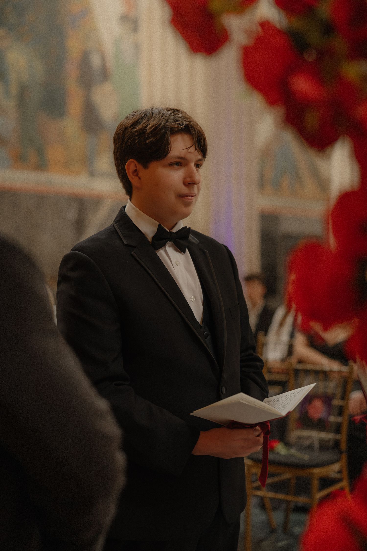 Young man in black tuxedo and bow tie holding papers at an elegant wedding ceremony with red floral decor.