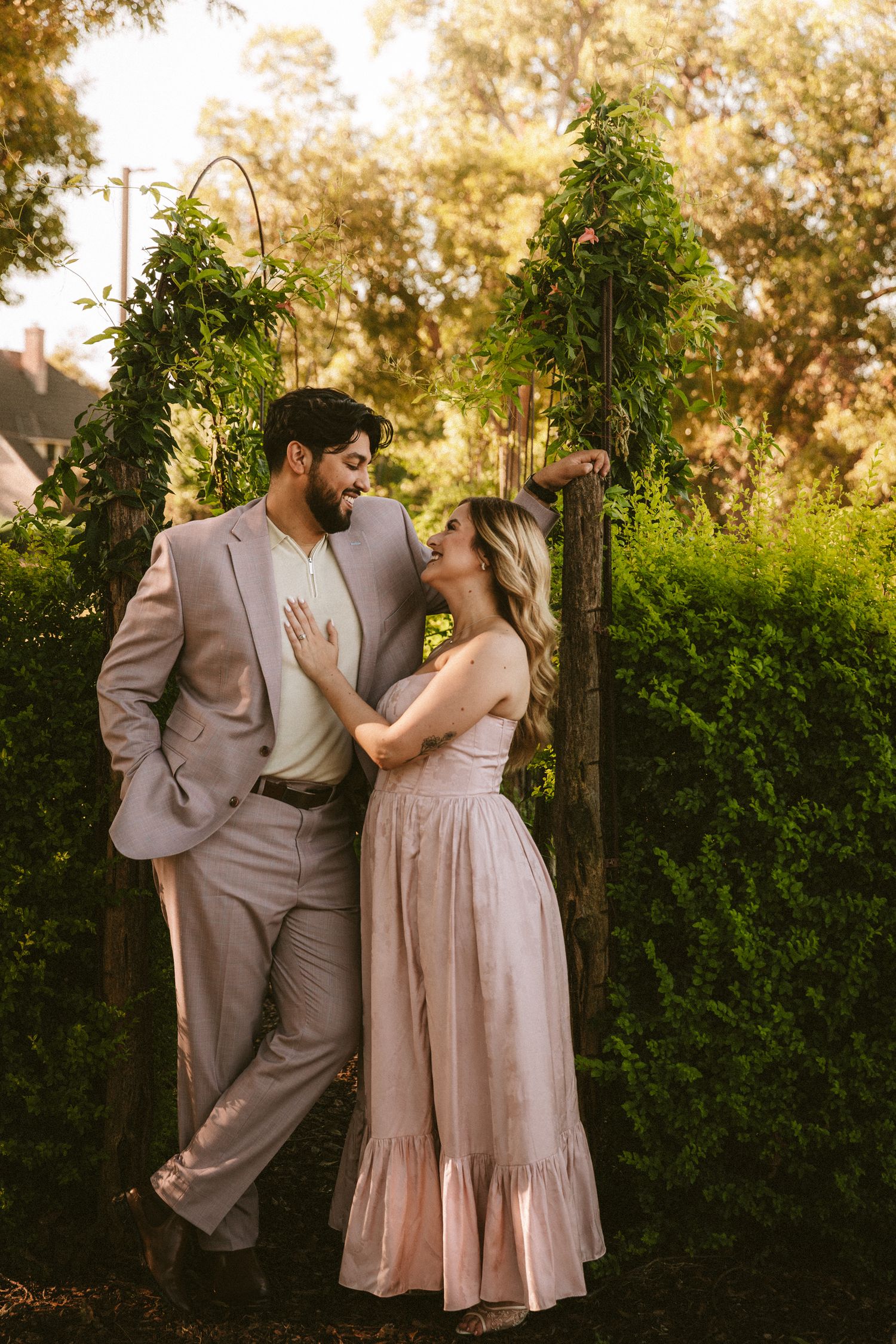 A romantic couple in blush-toned outfits share an intimate moment in a lush garden setting at golden hour.