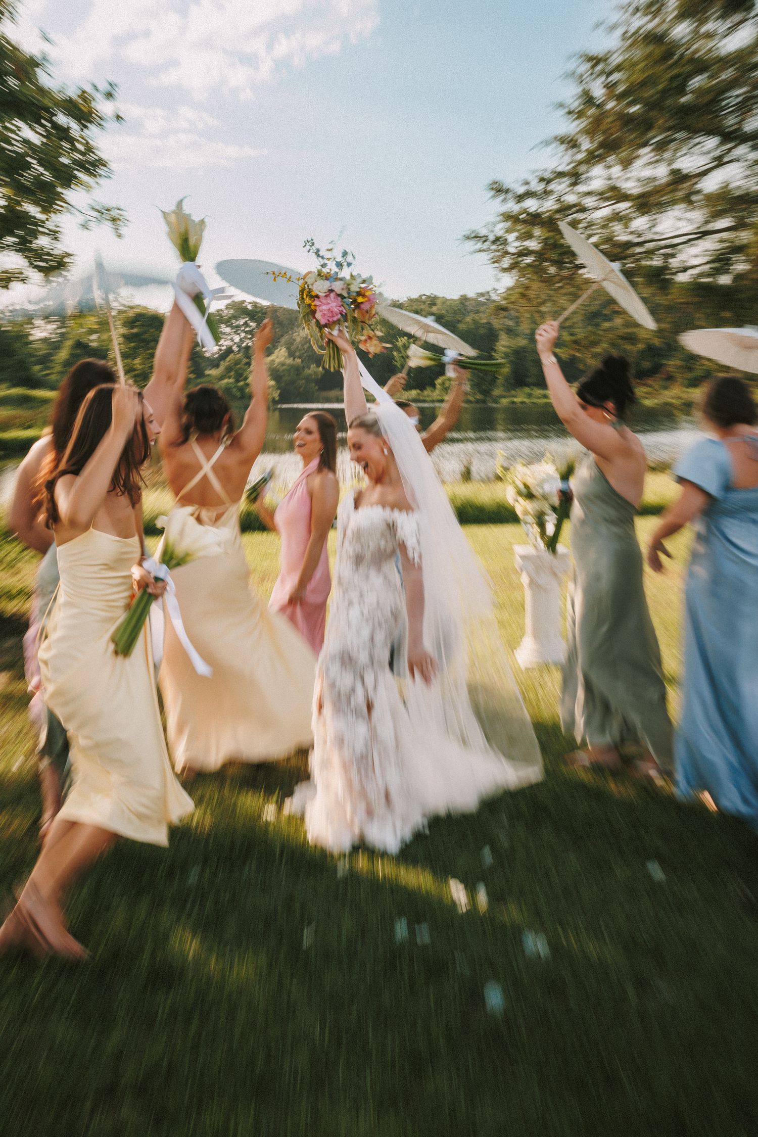 Bridesmaids in pastel dresses jumping and celebrating with bouquets on sunny lawn.