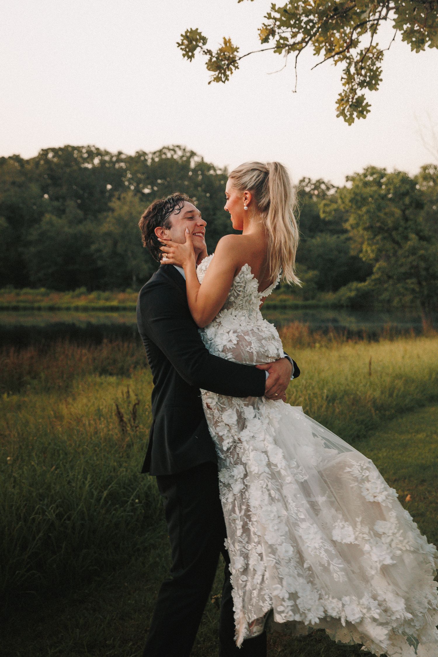 A groom lifts his bride in a lace wedding dress during sunset photos.