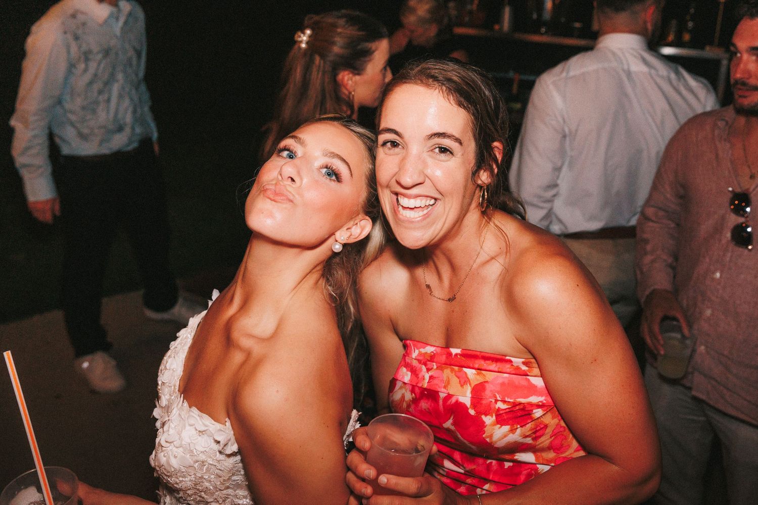 Two joyful friends pose together at a party, one wearing a white dress and one in a red floral dress.