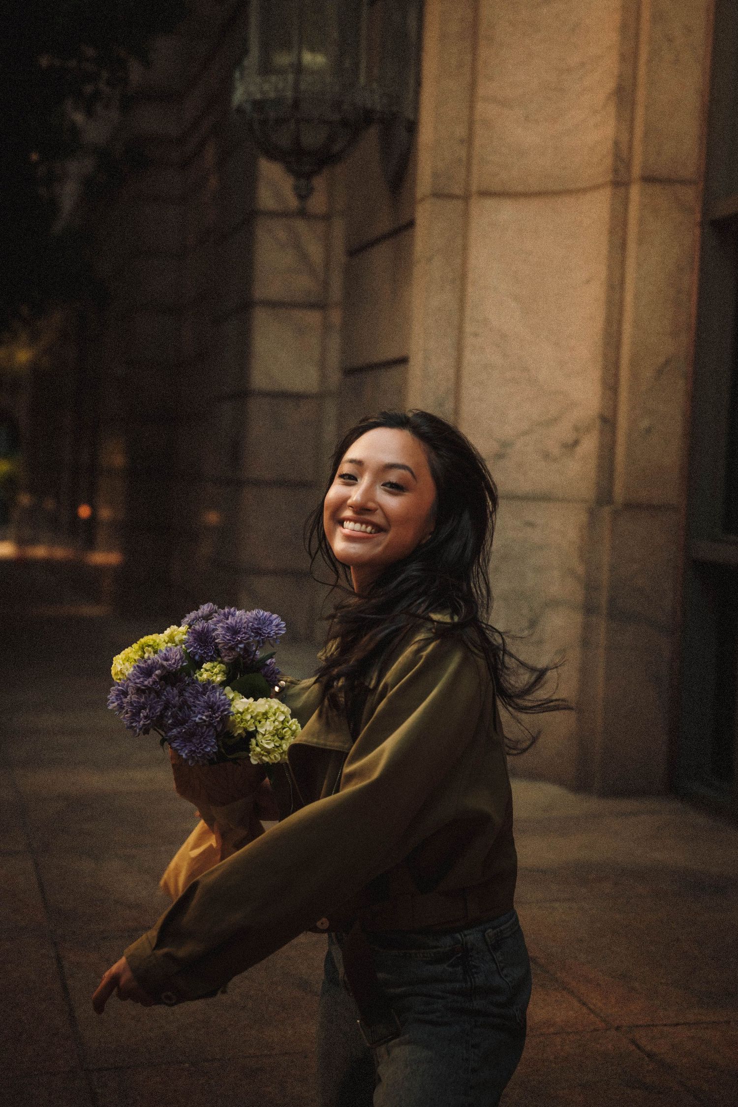 Woman in dark clothing spins joyfully while holding a colorful bouquet of flowers against stone architecture.