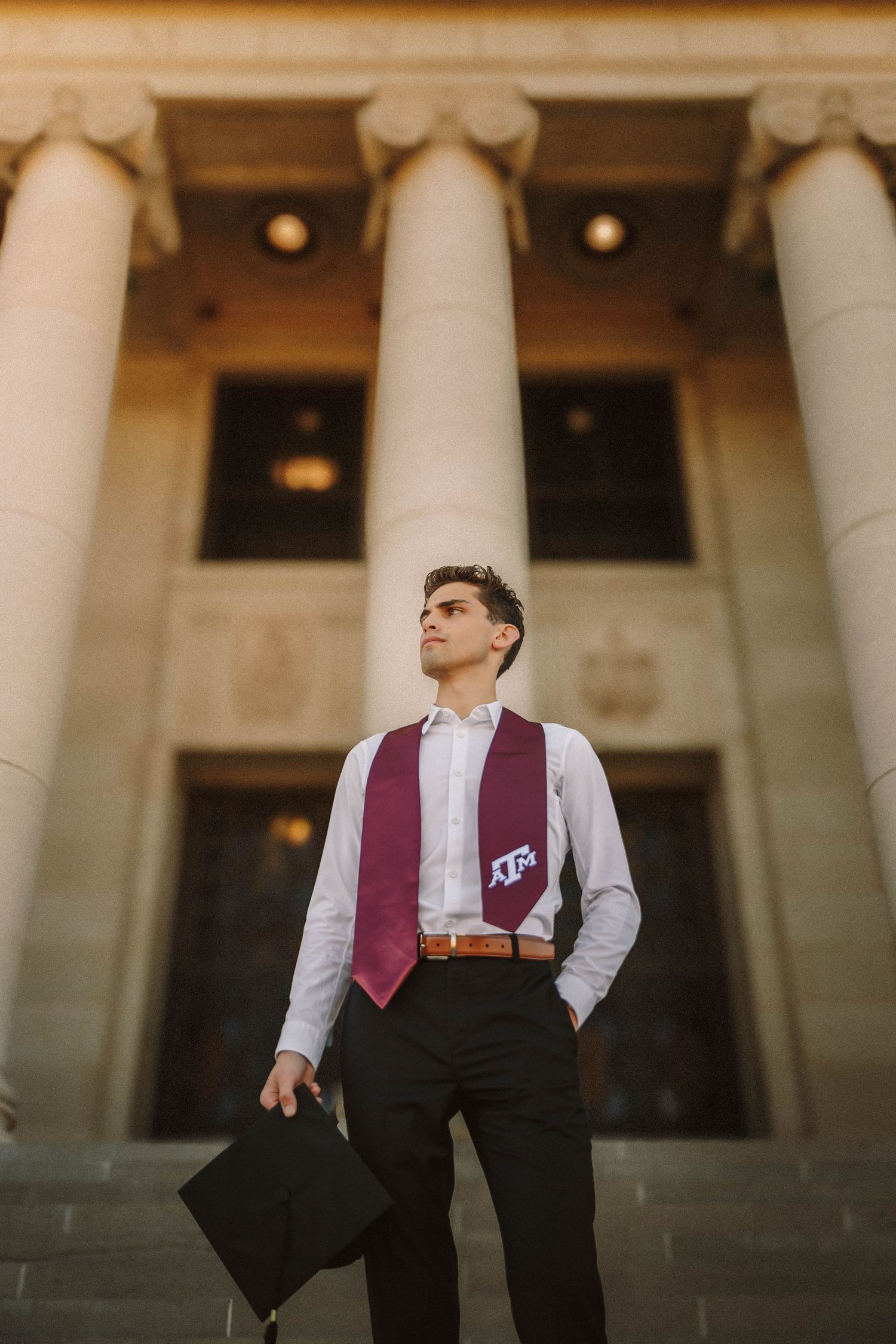 Graduate stands in front of classical columns in formal attire with burgundy tie looking up in contemplative pose.