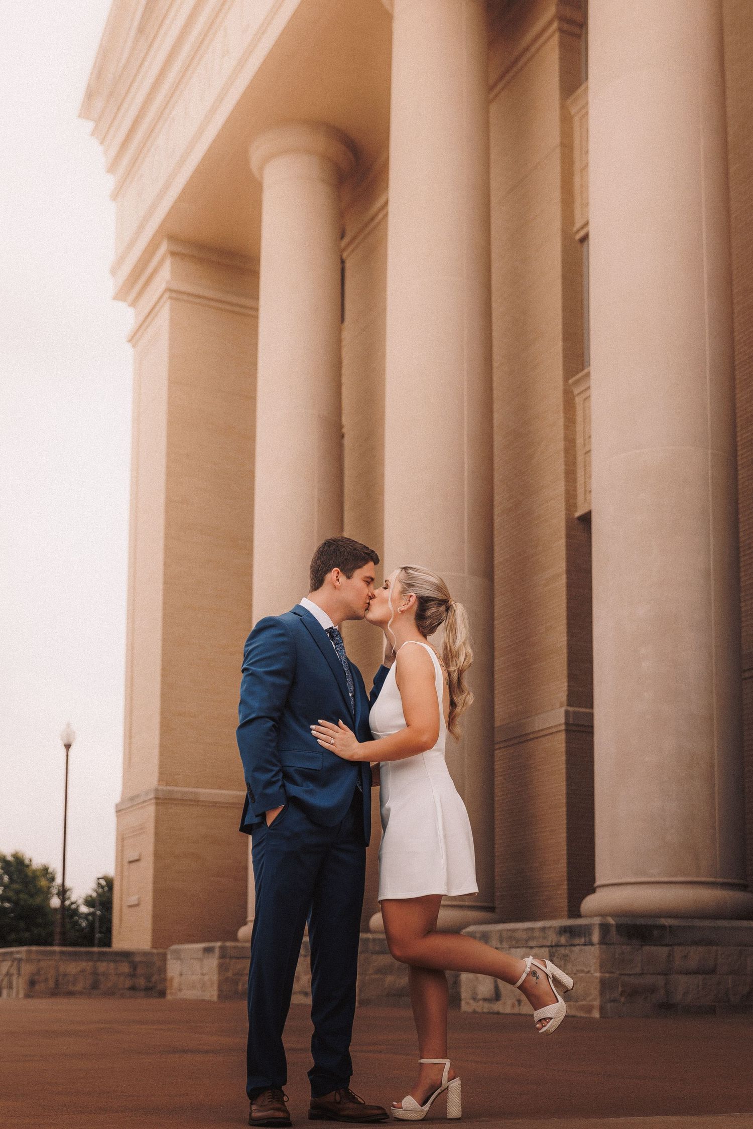 Romantic couple shares an intimate kiss in front of grand stone columns at dusk.