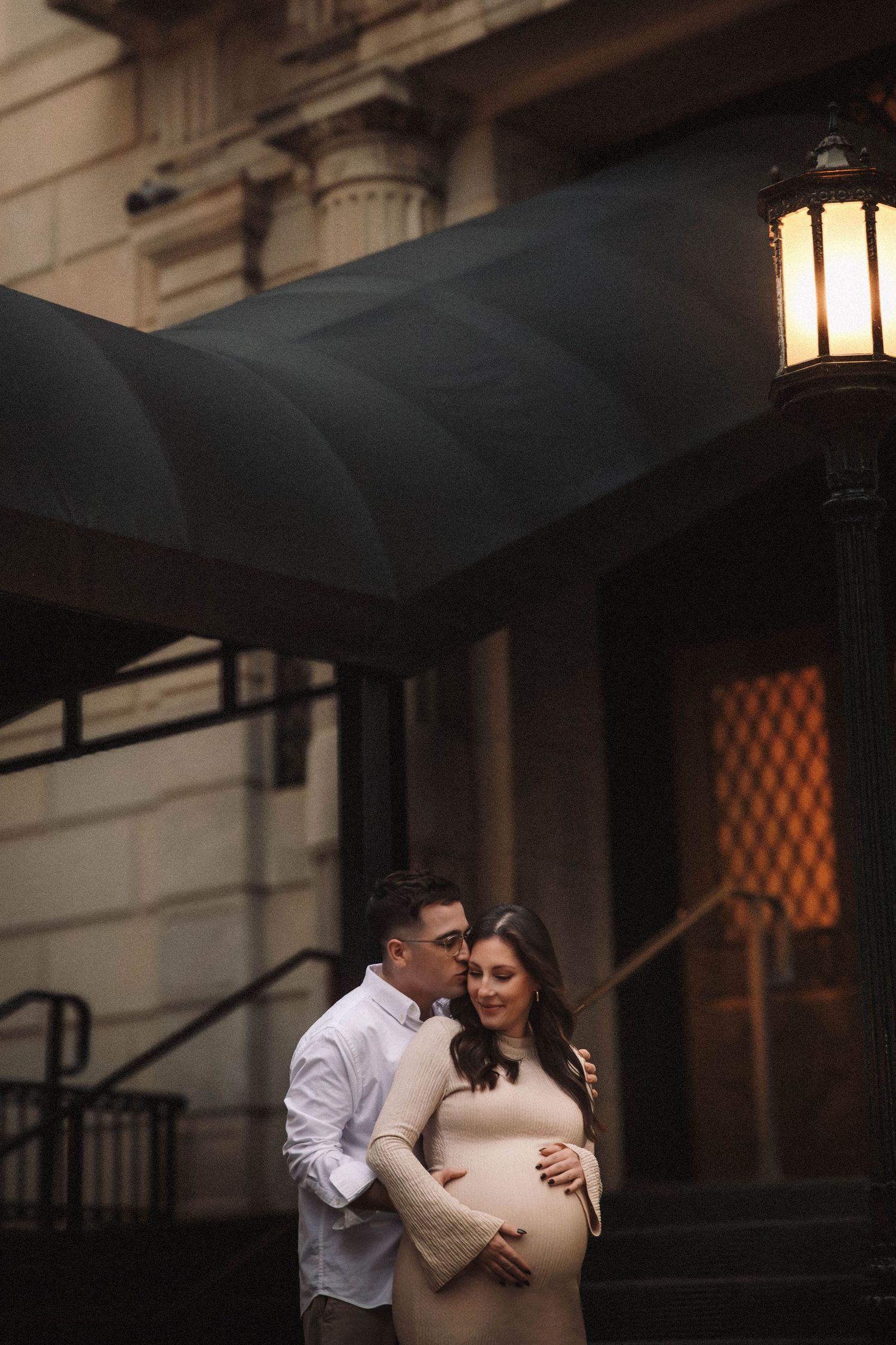 A romantic couple embraces on a dark evening outside a building with illuminated lanterns casting warm light.