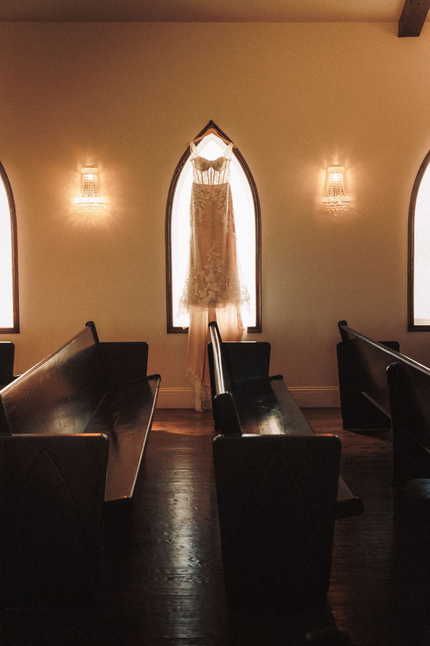 Lace wedding dress hanging in a chapel window between wooden pews and warm wall sconce lighting.