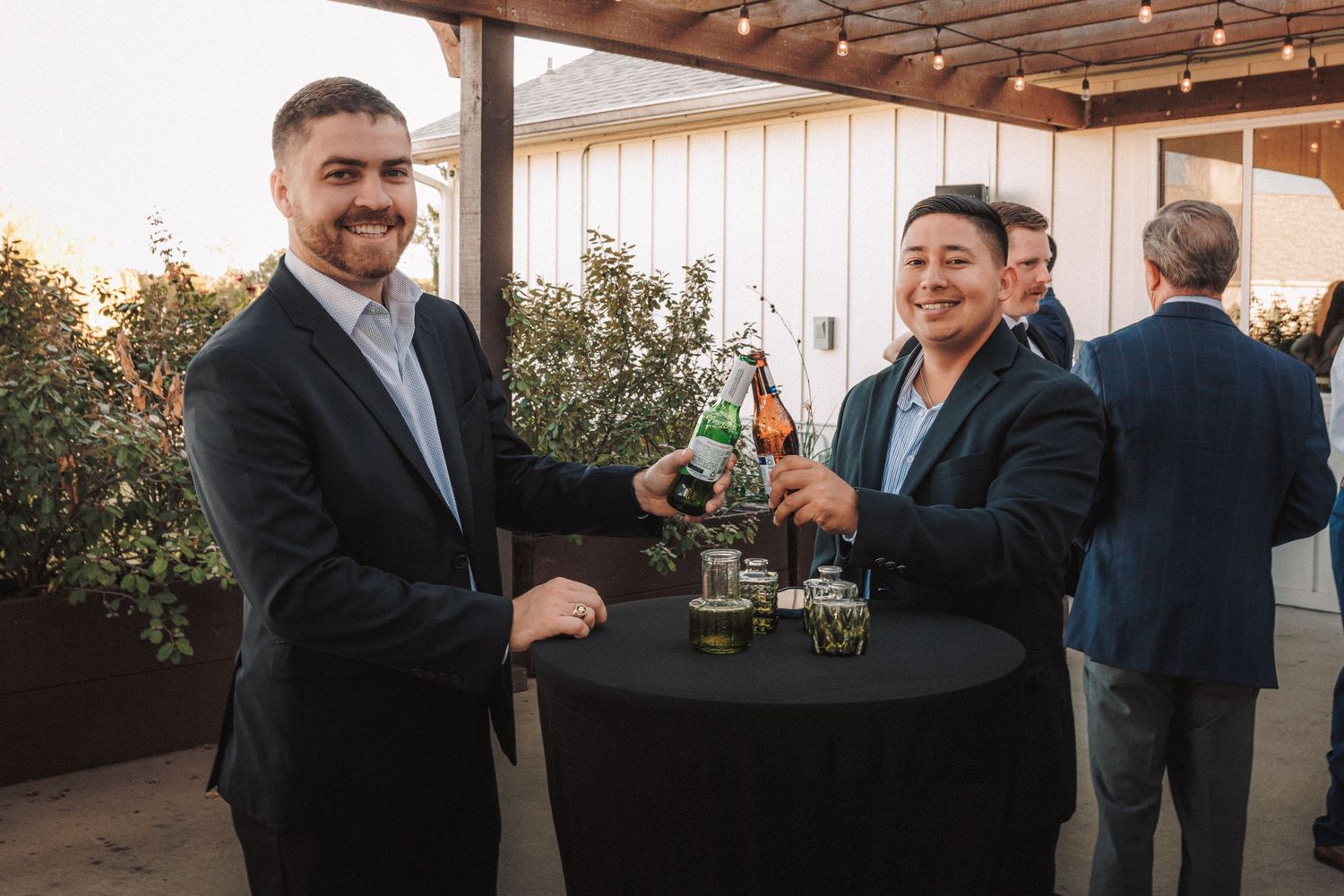 Two men in suits smile and clink beer bottles at a cocktail hour reception on an outdoor patio.
